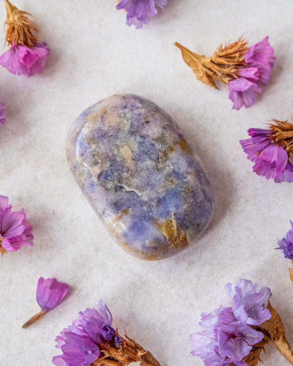 A polished lavender-blue tumbled stone crystal with natural white and gray marbling, arranged on a textured off-white surface and surrounded by dried purple flowers.