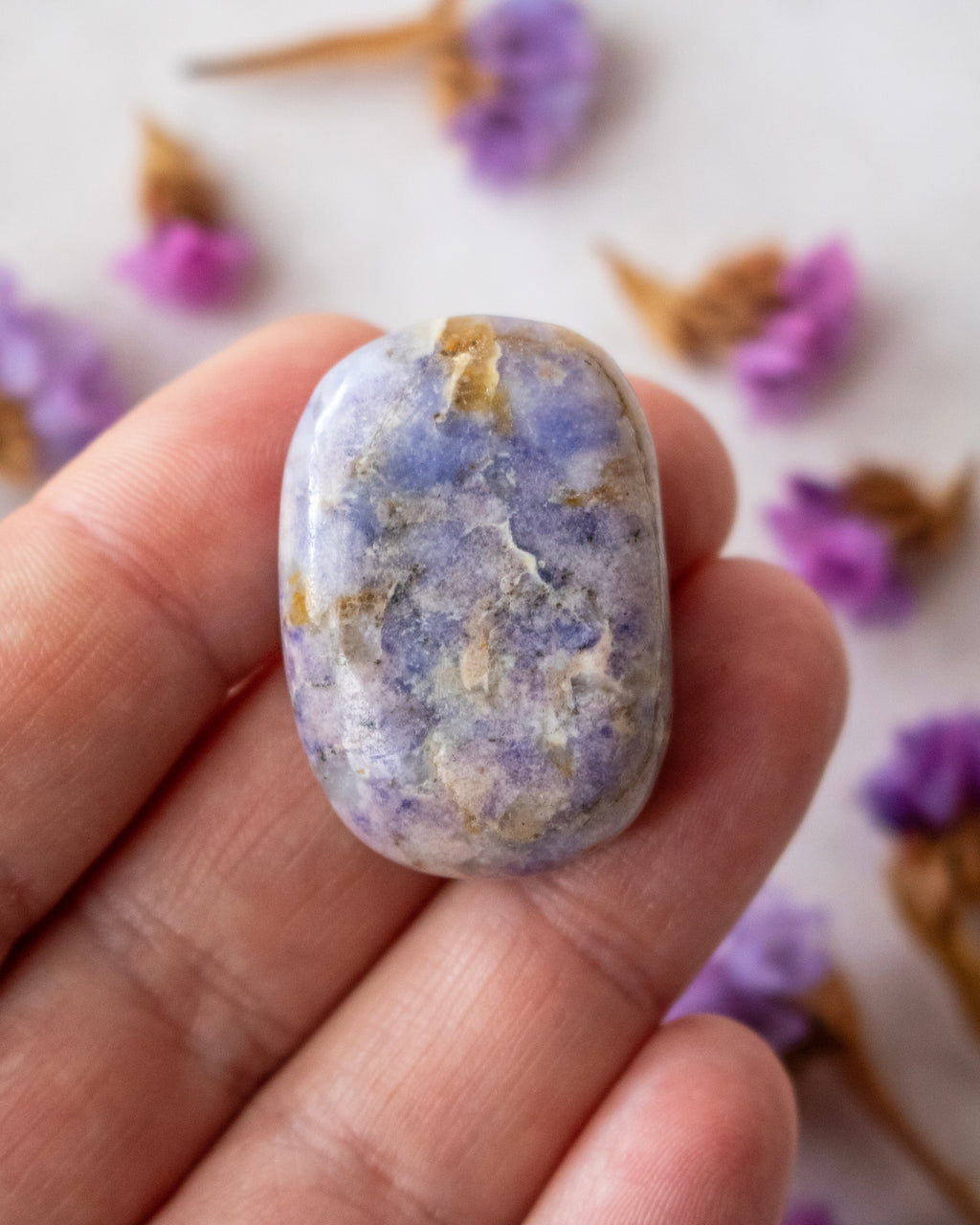 A polished lavender-blue tumbled stone crystal with natural white and gray marbling, held on someone's fingers and surrounded by dried purple flowers.