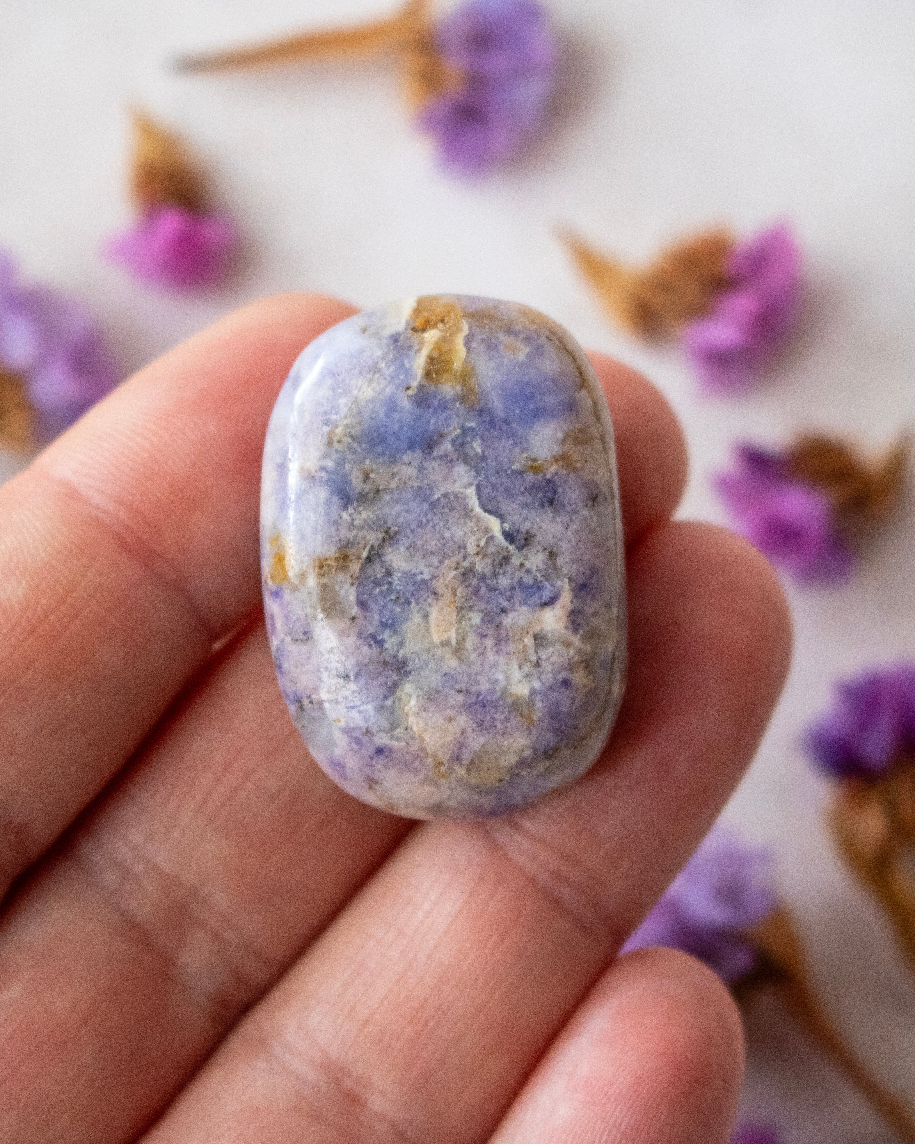 A polished lavender-blue tumbled stone crystal with natural white and gray marbling, held on someone's fingers and surrounded by dried purple flowers.