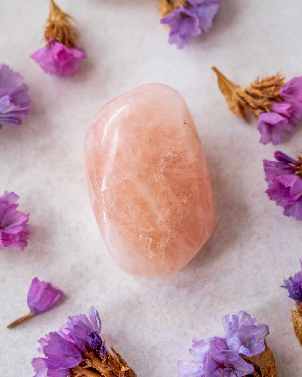 A smooth, polished pink crystal stone centered on a light textured surface, surrounded by scattered dried purple flowers.