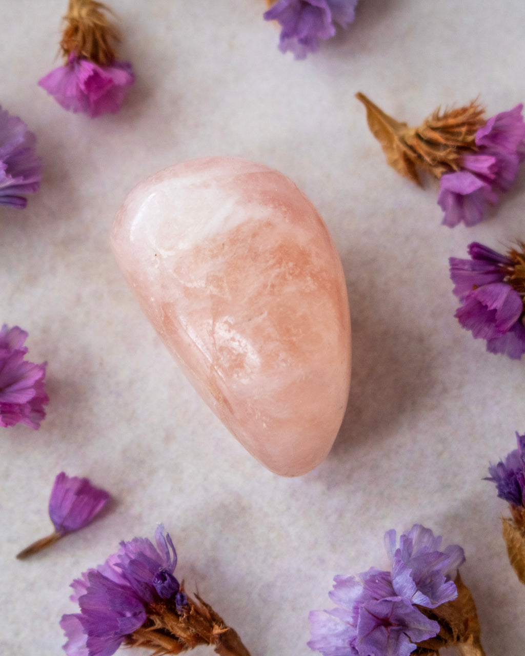 A polished pale pink crystal stone centered on a light neutral surface, surrounded by scattered dried purple flowers.