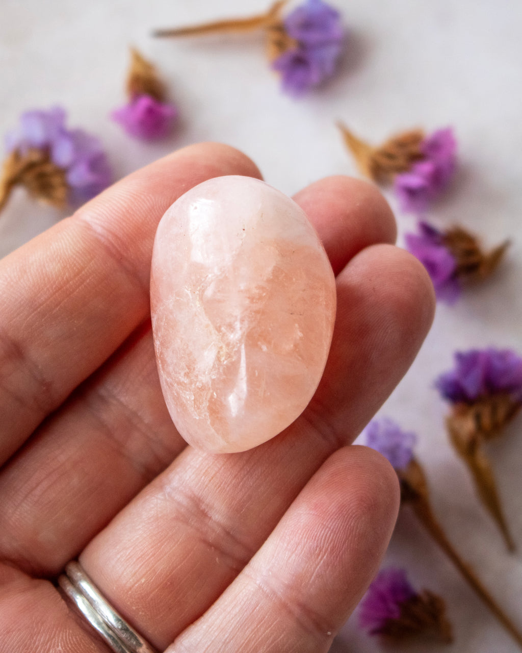 A polished peach-pink crystal held in a person's hand, with dried purple flowers scattered in the background on a light surface.