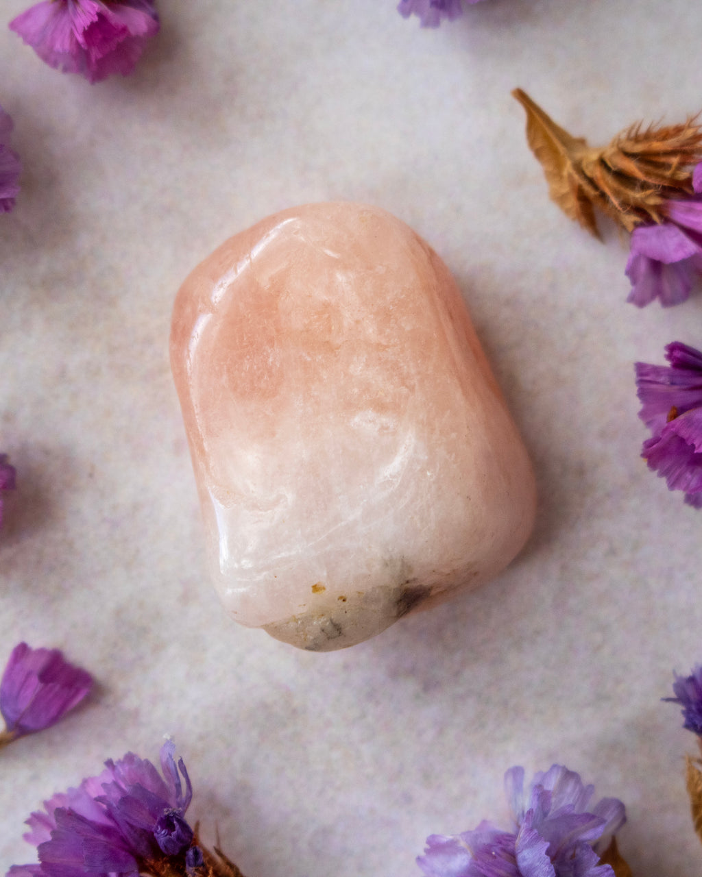 A smooth, polished peach-colored tumbled stone crystal resting on a textured light gray surface, surrounded by scattered purple dried flowers.