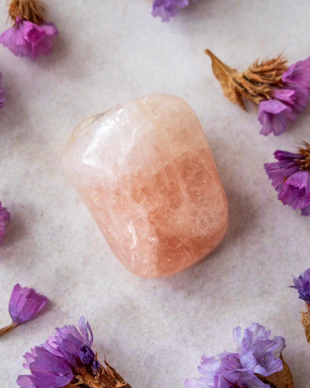 A polished peach-colored tumbled crystal stone with a smooth, rounded shape, resting on a textured off-white surface and surrounded by scattered dried purple flowers.