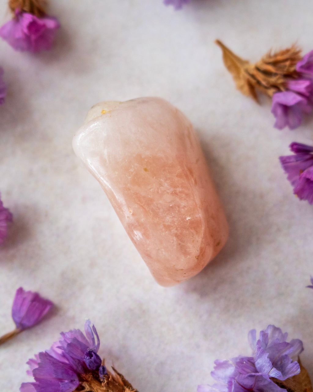 A polished crystal stone with a smooth, rounded shape, resting on a light textured surface and surrounded by scattered dried purple flowers.