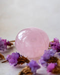 a round pink rose quartz polished crystal surrounded by purple dried flowers on a off-white background