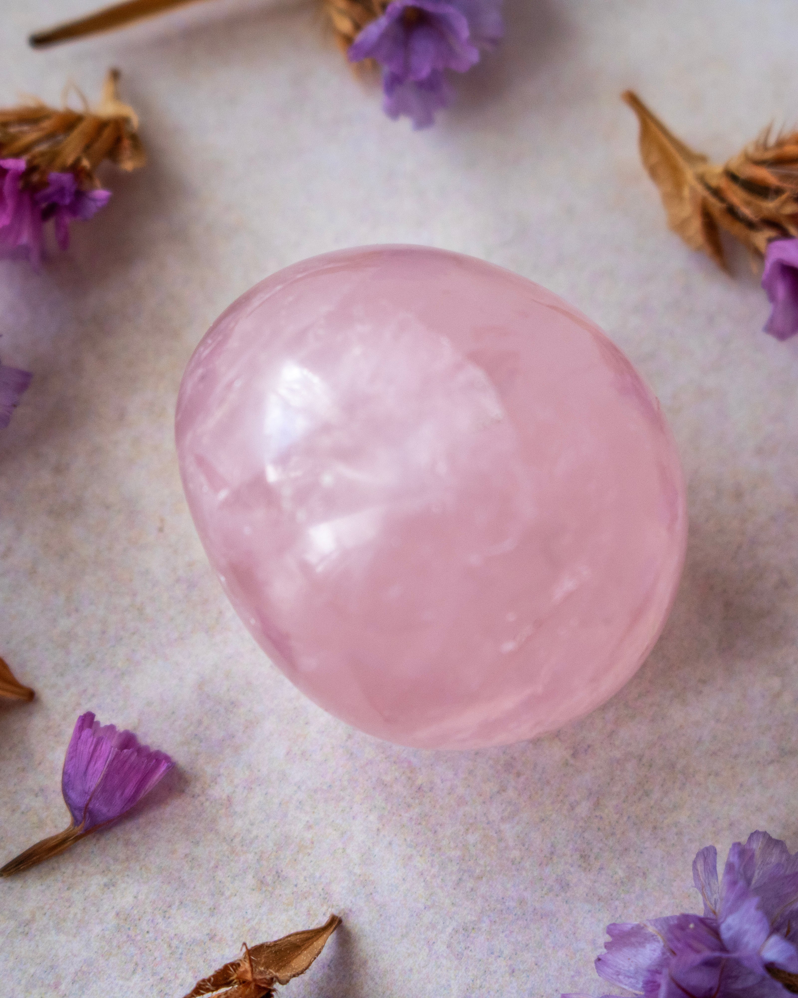 A smooth, polished pink crystal egg resting on a textured light beige surface, surrounded by scattered dried brown petals and small purple flowers.