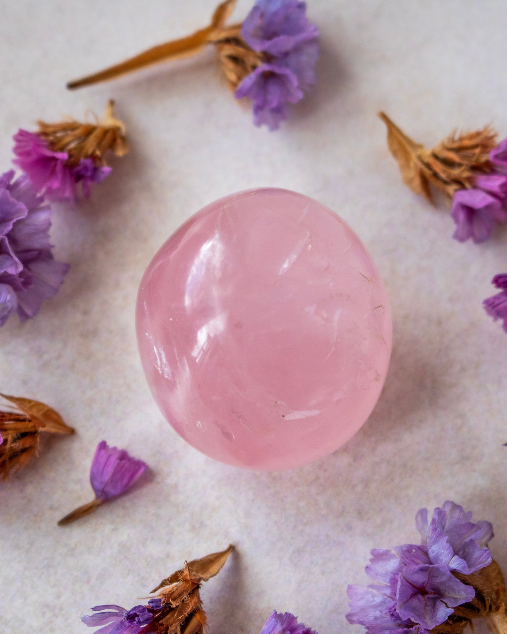 A smooth, polished pink rose quartz crystal ball centered on a textured off-white surface, surrounded by scattered dried purple flowers.