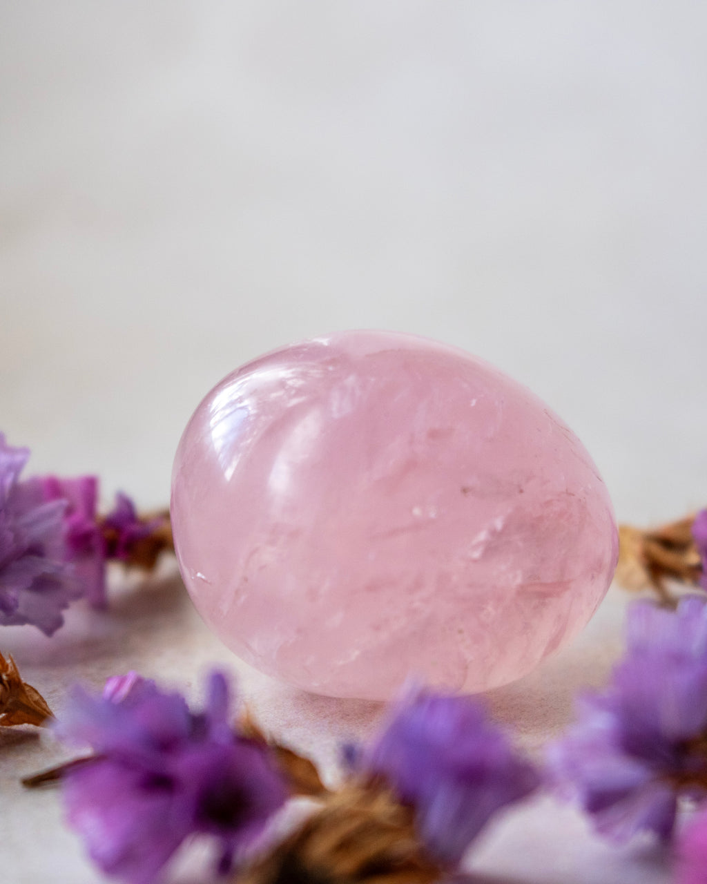 A polished pink rose quartz crystal sphere resting on a light surface, surrounded by small purple flowers.