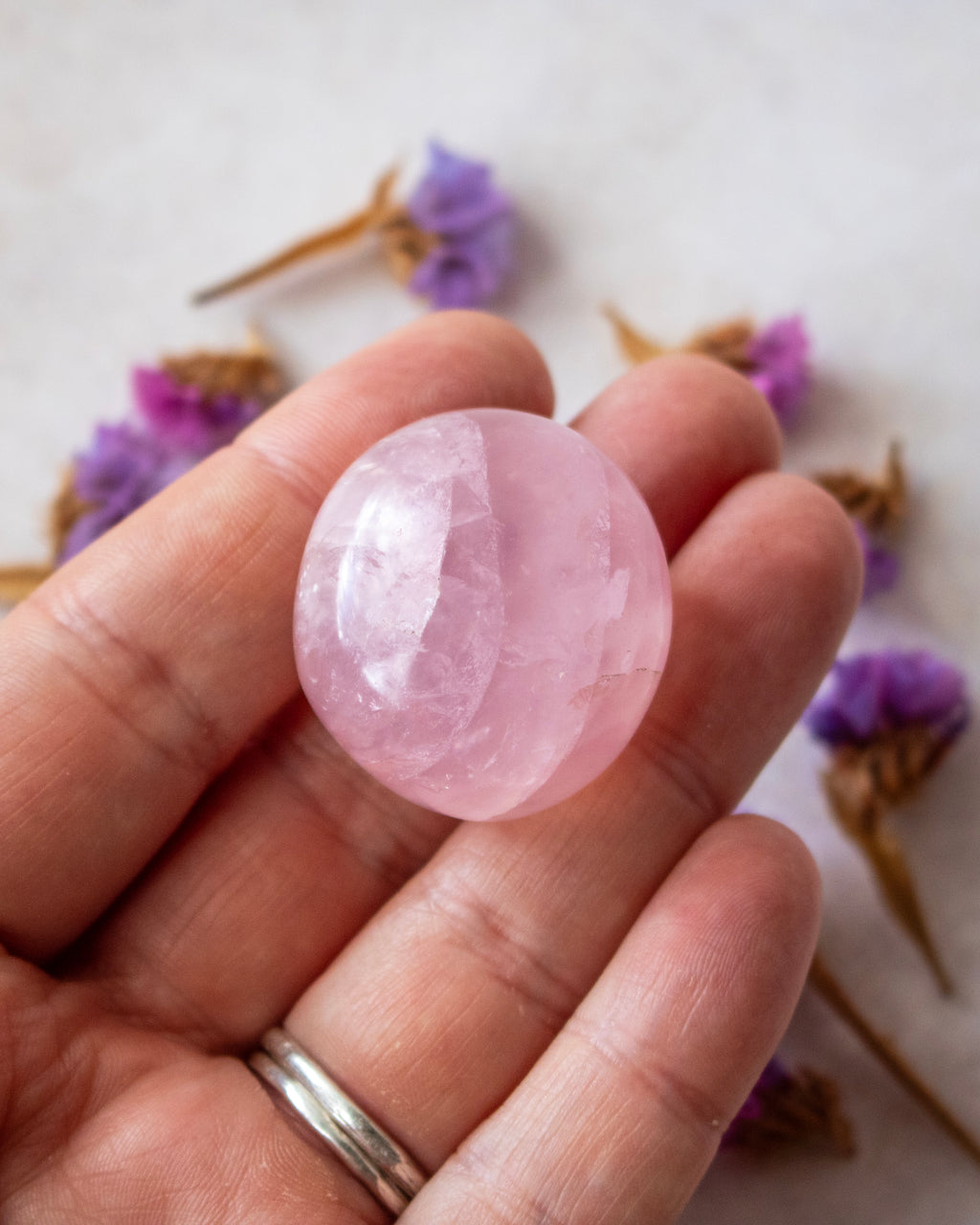 A hand holding a polished pink rose quartz gemstone, with dried purple flowers blurred in the background on a light surface.