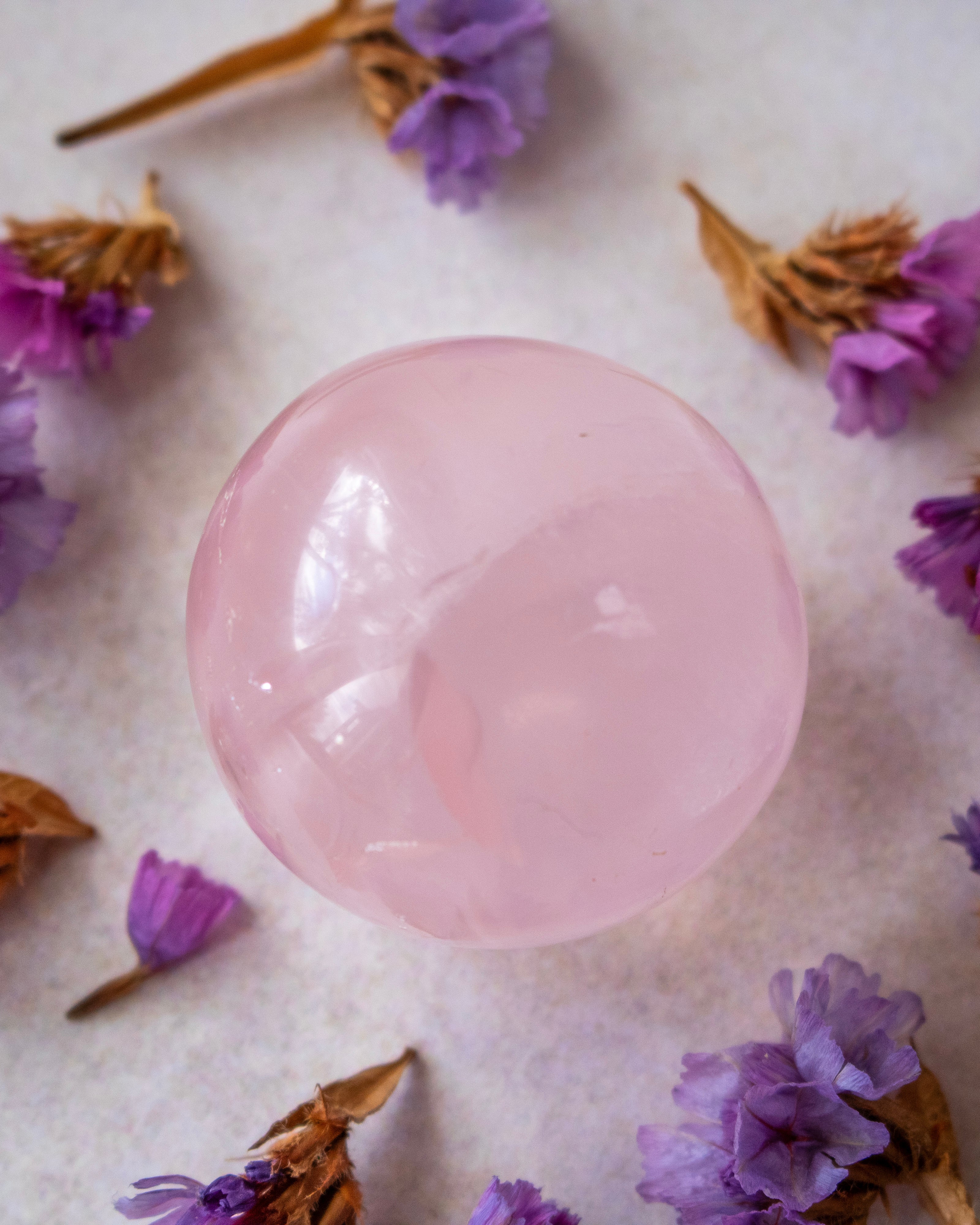 A smooth, polished pink crystal sphere resting on a textured off-white surface, surrounded by scattered dried purple flowers.