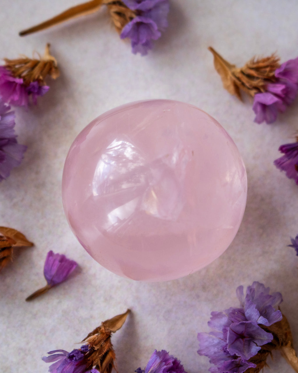 A smooth, polished light pink crystal sphere centered on a textured off-white surface, surrounded by scattered dried purple flowers.