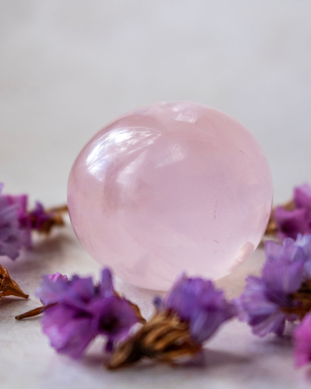 A smooth, polished light pink crystal sphere resting on a white surface and surrounded by small purple dried flowers.