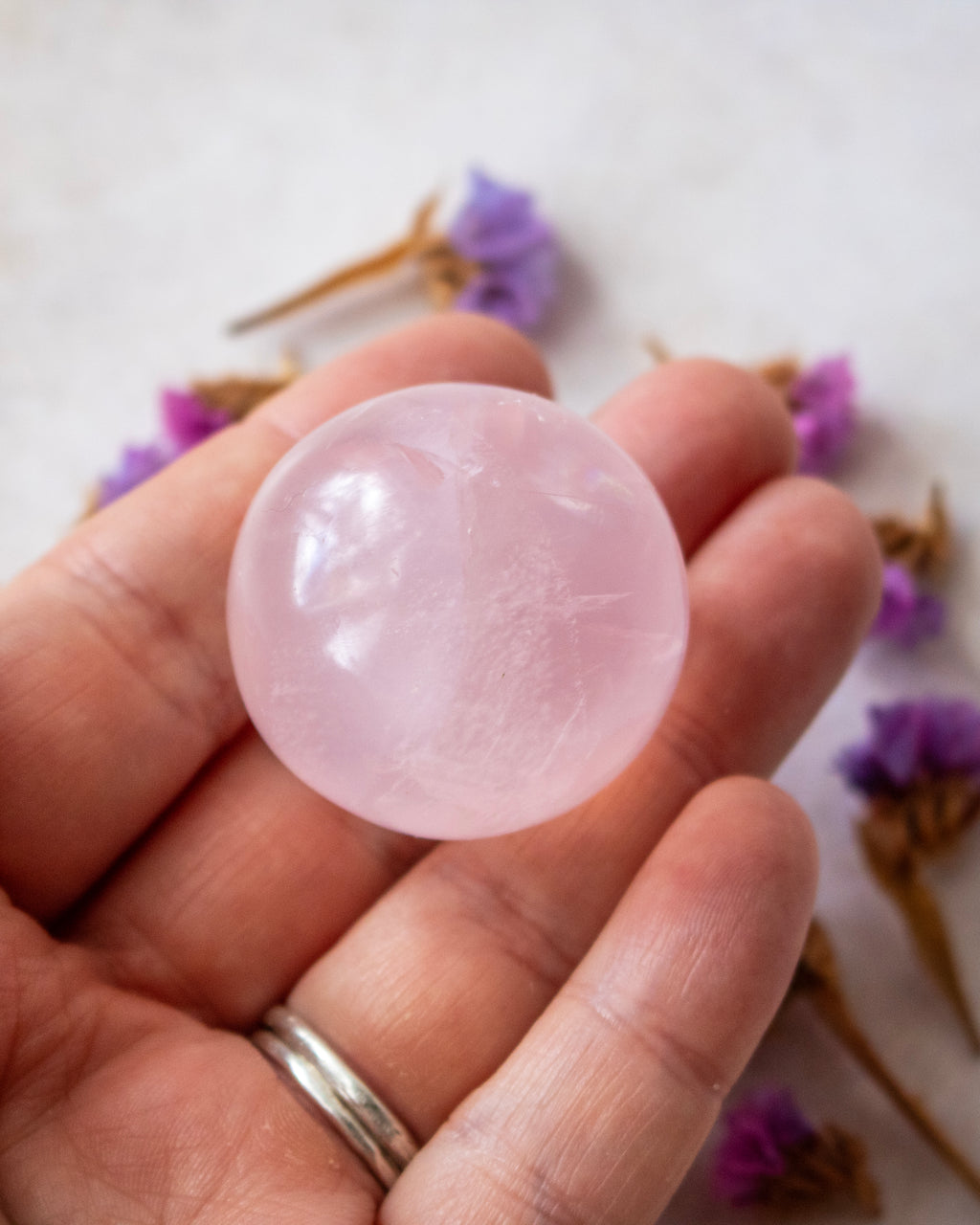 A hand holding a smooth, polished pink stone orb, with dried purple flowers blurred in the background.