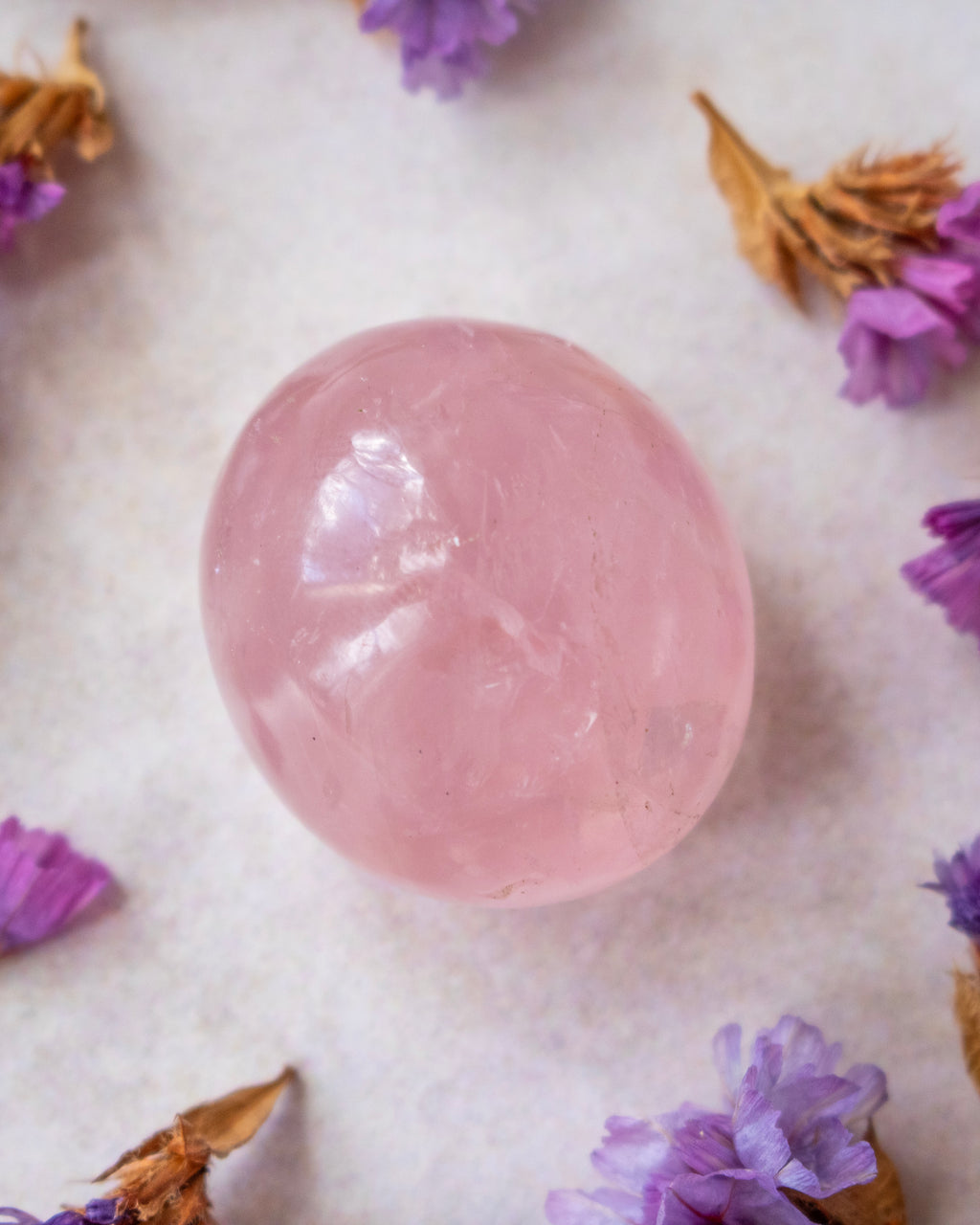 A smooth, polished pink quartz crystal sphere resting on a textured off-white surface, surrounded by scattered dried lavender and brown flower petals.