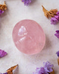 A smooth, polished pink quartz crystal sphere resting on a textured off-white surface, surrounded by scattered dried lavender and brown flower petals.