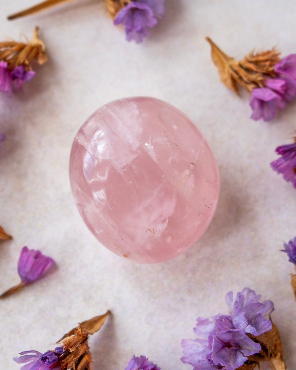 A smooth, polished pink crystal sphere resting on a textured off-white surface, surrounded by scattered dried purple flowers.