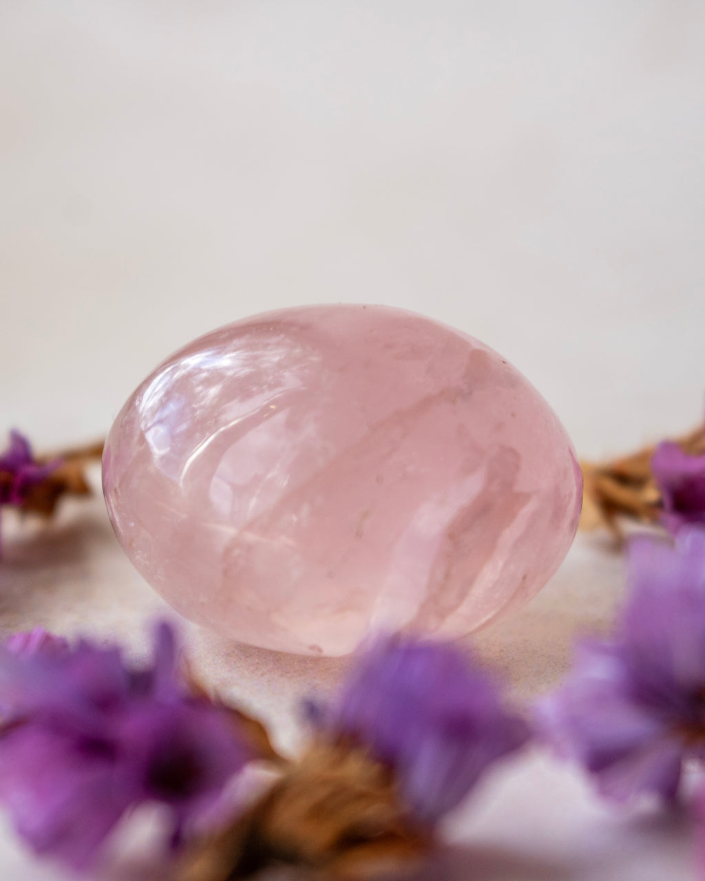 A polished pink rose quartz crystal ball resting on a light surface, surrounded by blurred purple flowers.