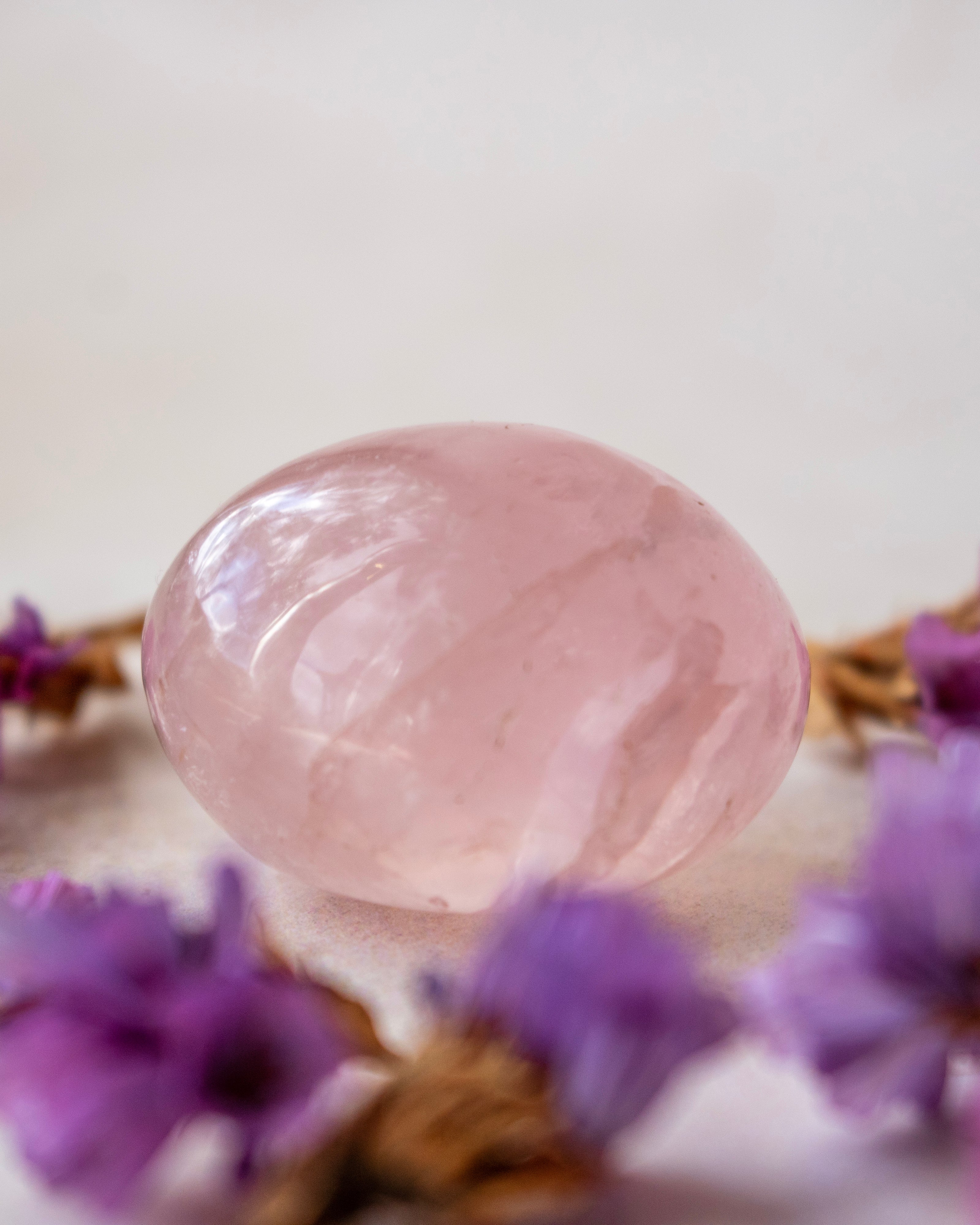 A polished pink rose quartz crystal ball resting on a light surface, surrounded by blurred purple flowers.