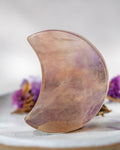 A polished crescent-shaped crystal tool, likely quartz, with natural lavender and peach zoned patterns, standing upright on a white marble surface with blurred purple flowers in the background.