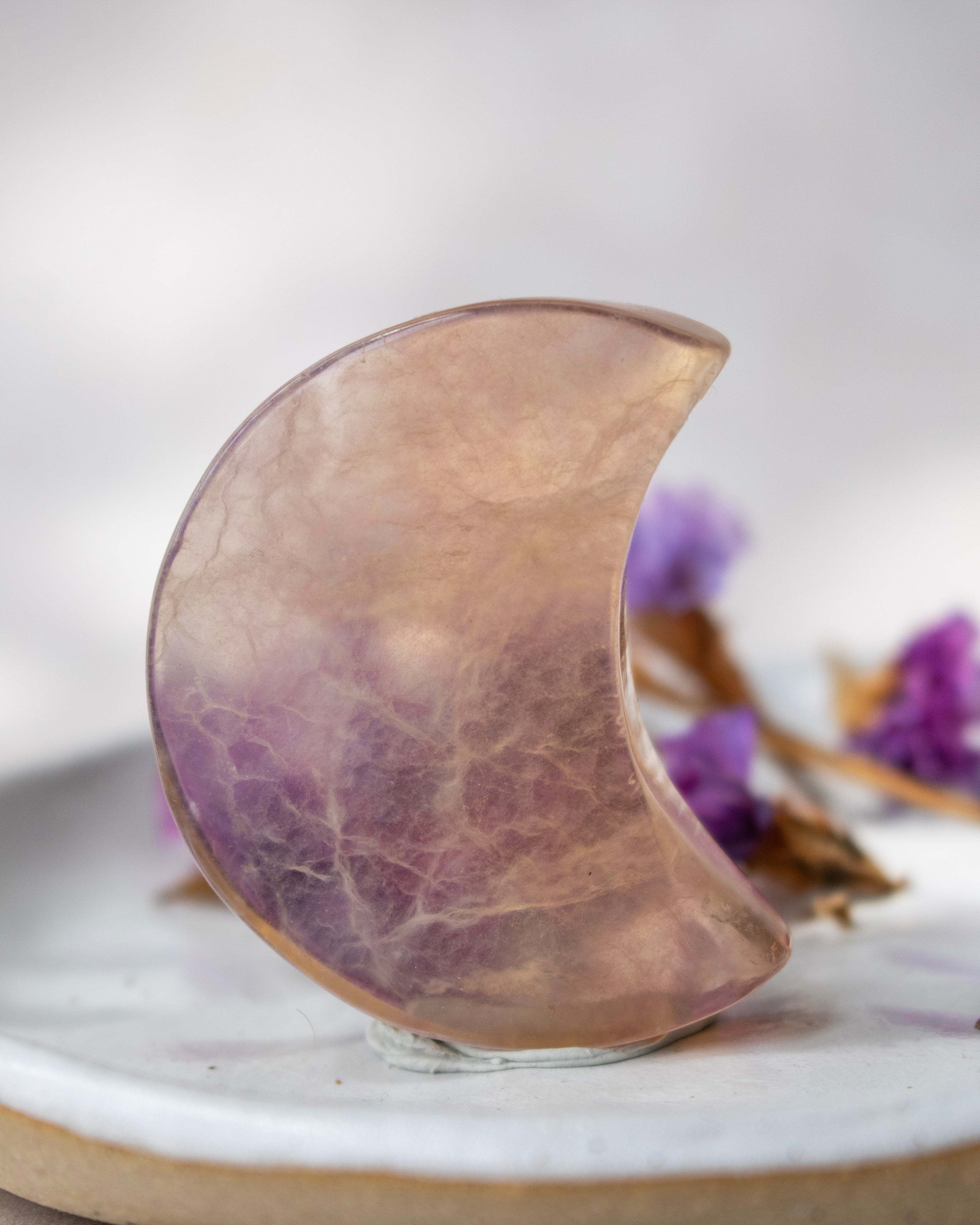 A crescent moon-shaped polished stone object with a marbled purple and beige pattern, displayed on a small clear stand against a soft blurred background with dried purple flowers.