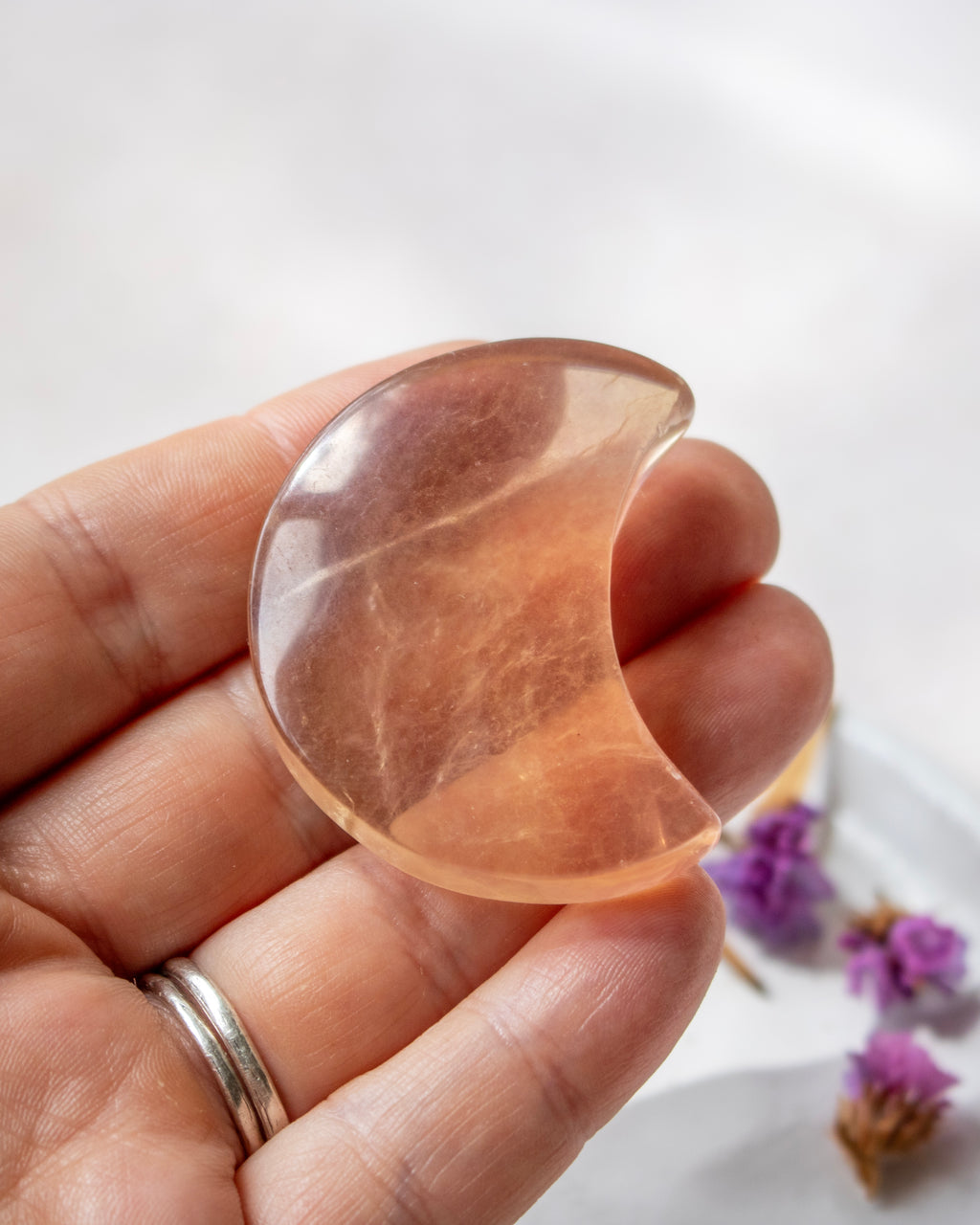 A hand holds a polished crescent moon-shaped crystal pendant made of peachy orange stone, with dried purple flowers on a soft white surface in the background.