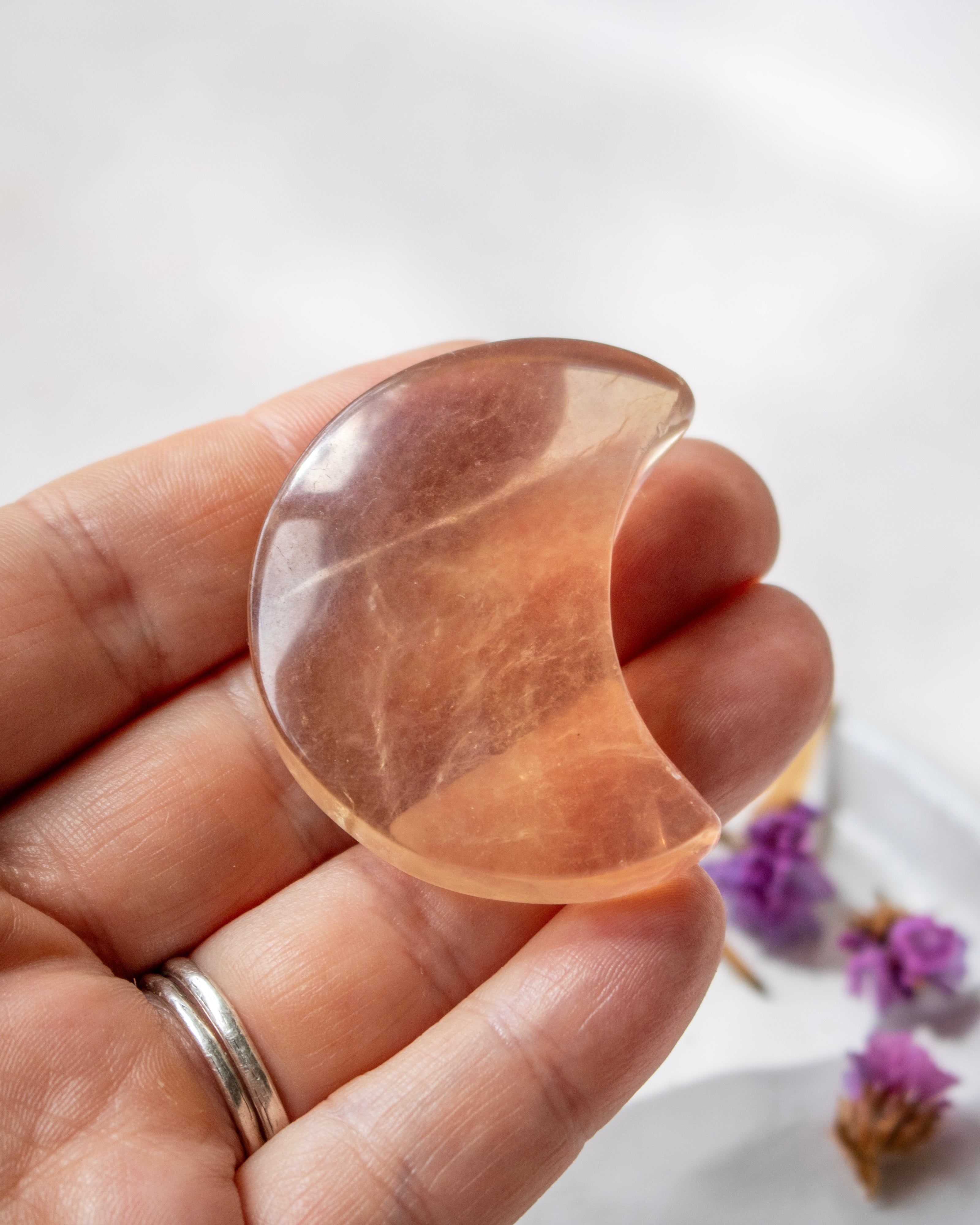 A hand holds a polished crescent moon-shaped crystal pendant made of peachy orange stone, with dried purple flowers on a soft white surface in the background.