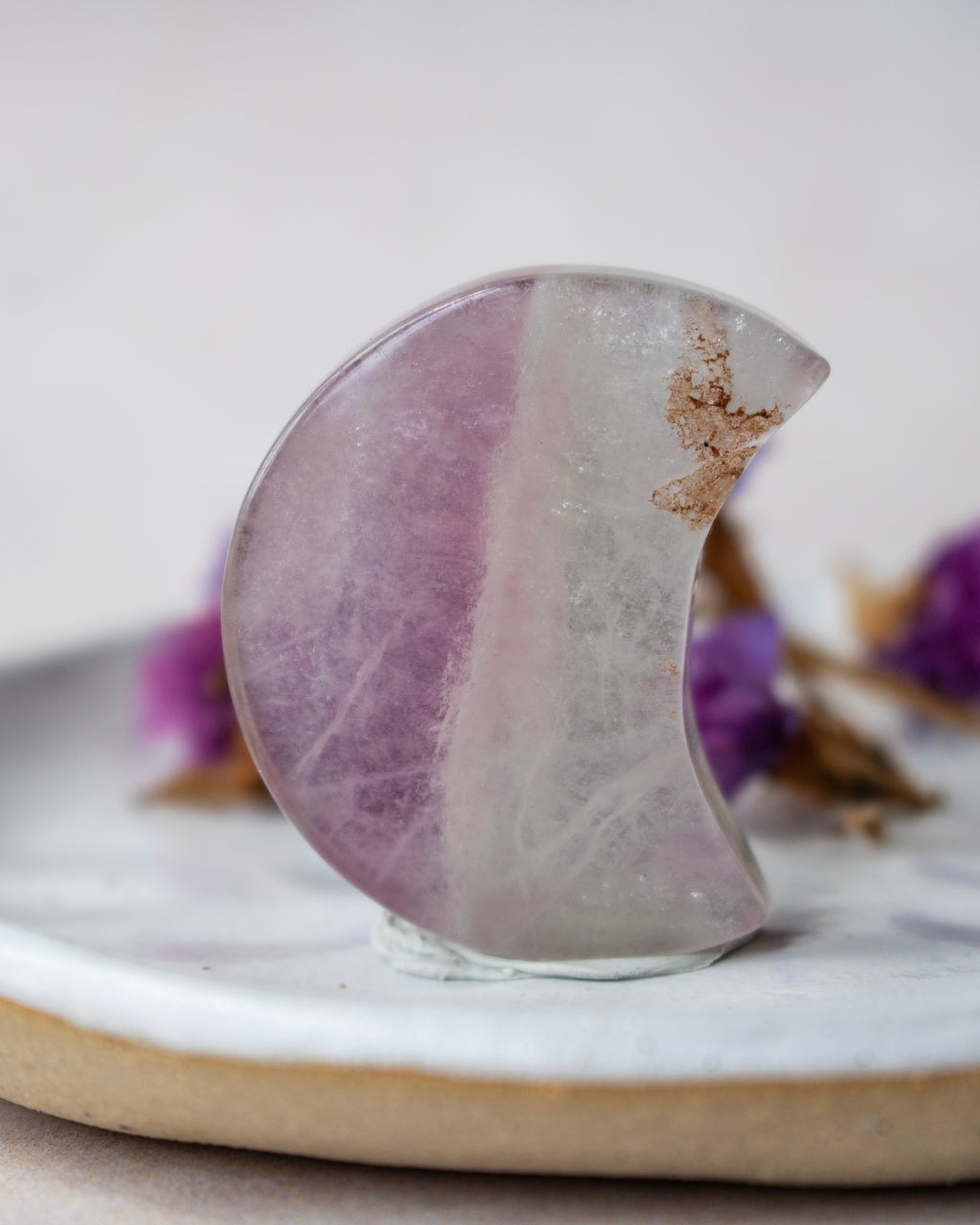 A polished crescent moon-shaped crystal with lavender purple and milky white bands, resting on a white ceramic dish with dried purple flowers in the background.
