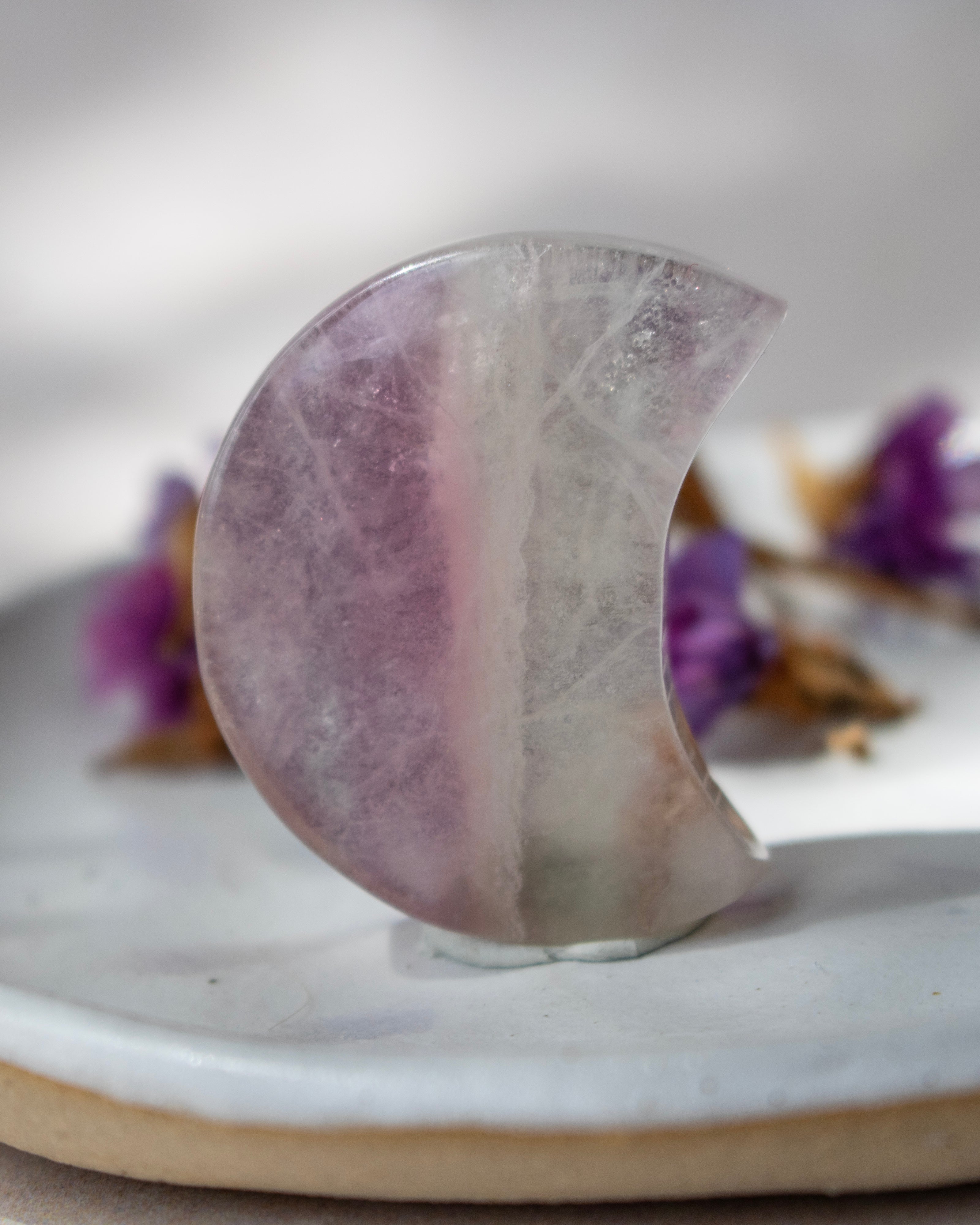 A crescent-shaped polished crystal stone with natural purple and milky white marbling, displayed on a light gray ceramic dish with blurred purple flowers in the background.