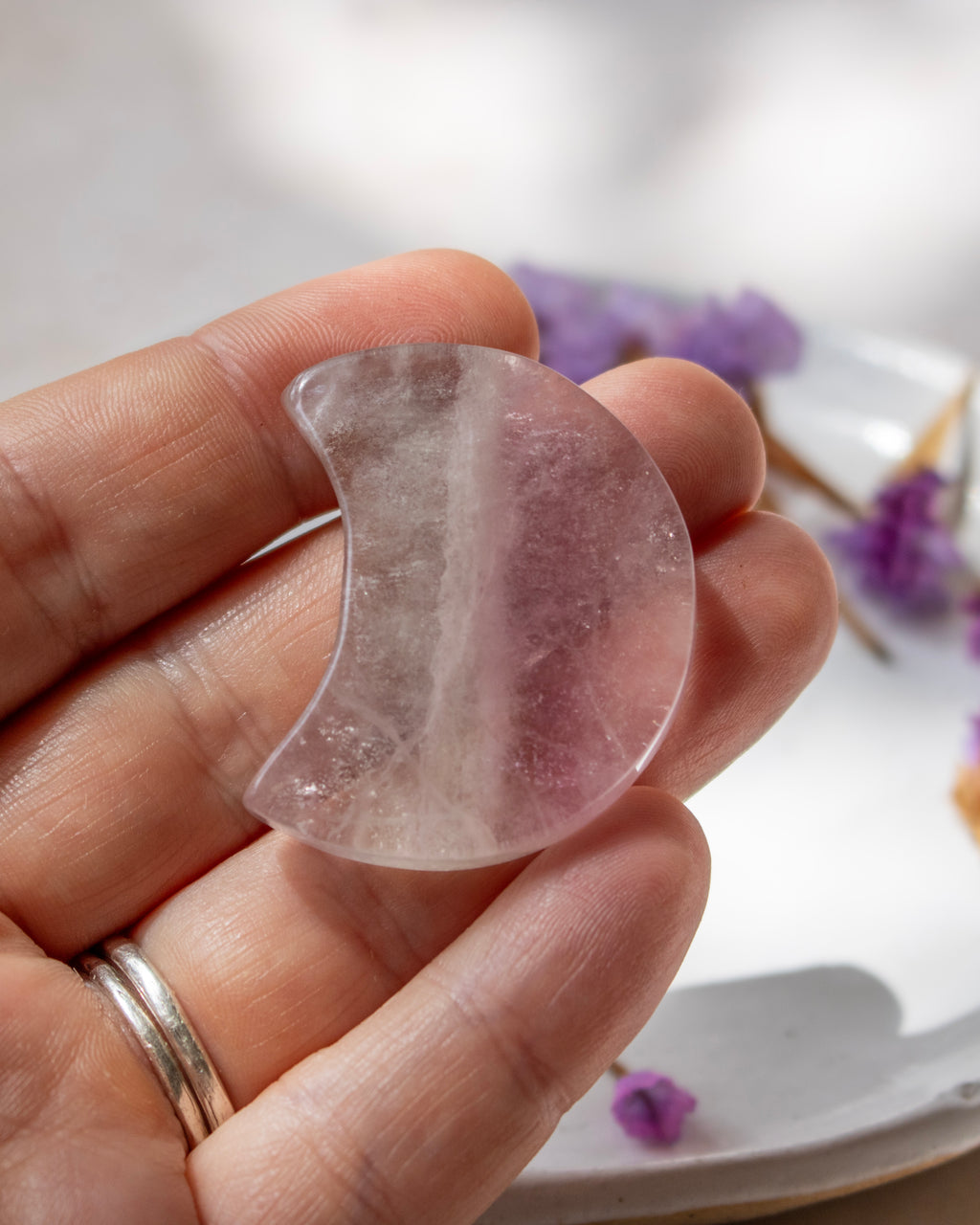 A hand holding a crescent-shaped translucent pink stone or crystal against a softly blurred white background with small purple flowers.