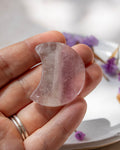 A hand holding a crescent-shaped translucent pink stone or crystal against a softly blurred white background with small purple flowers.