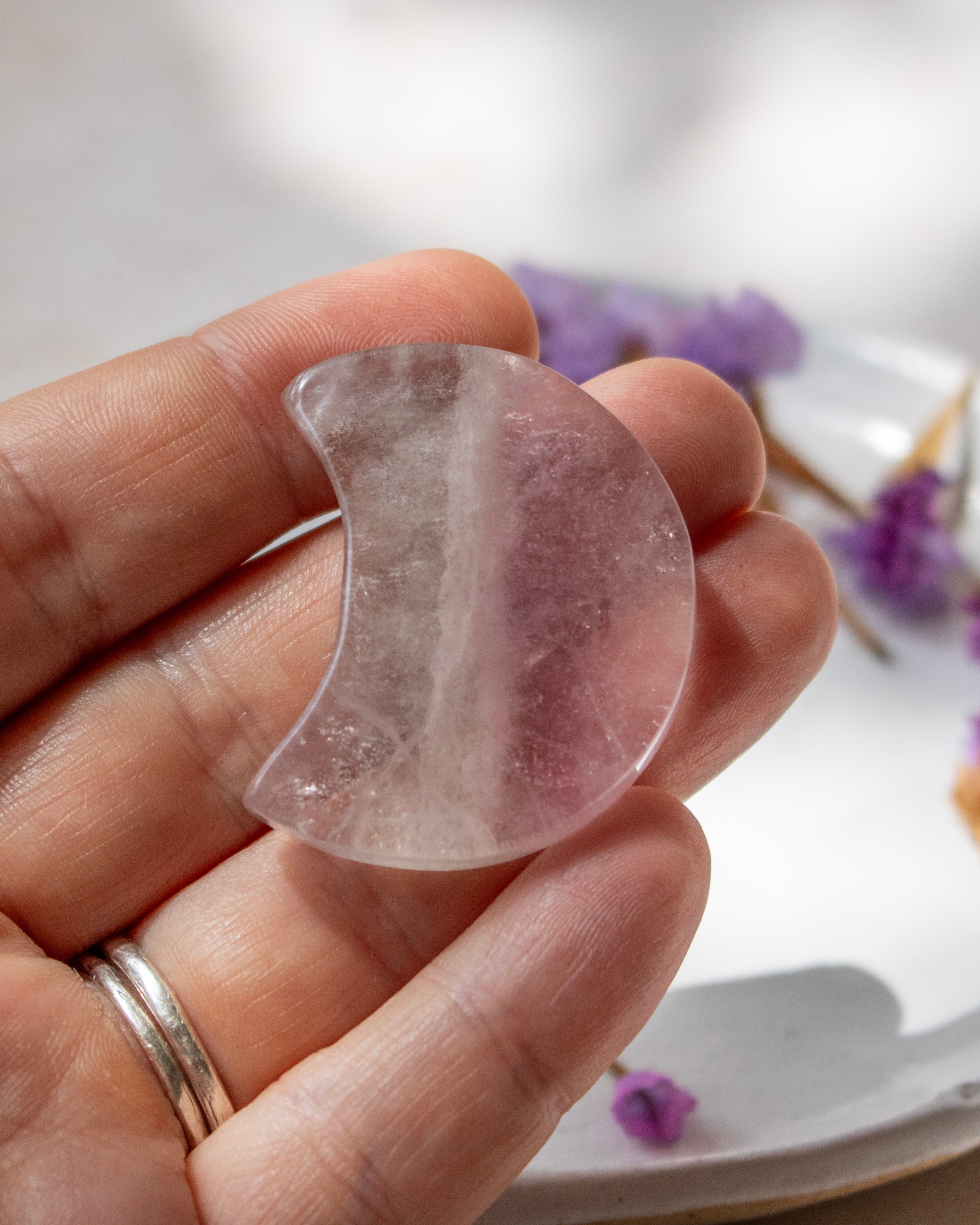 A hand holding a crescent-shaped translucent pink stone or crystal against a softly blurred white background with small purple flowers.