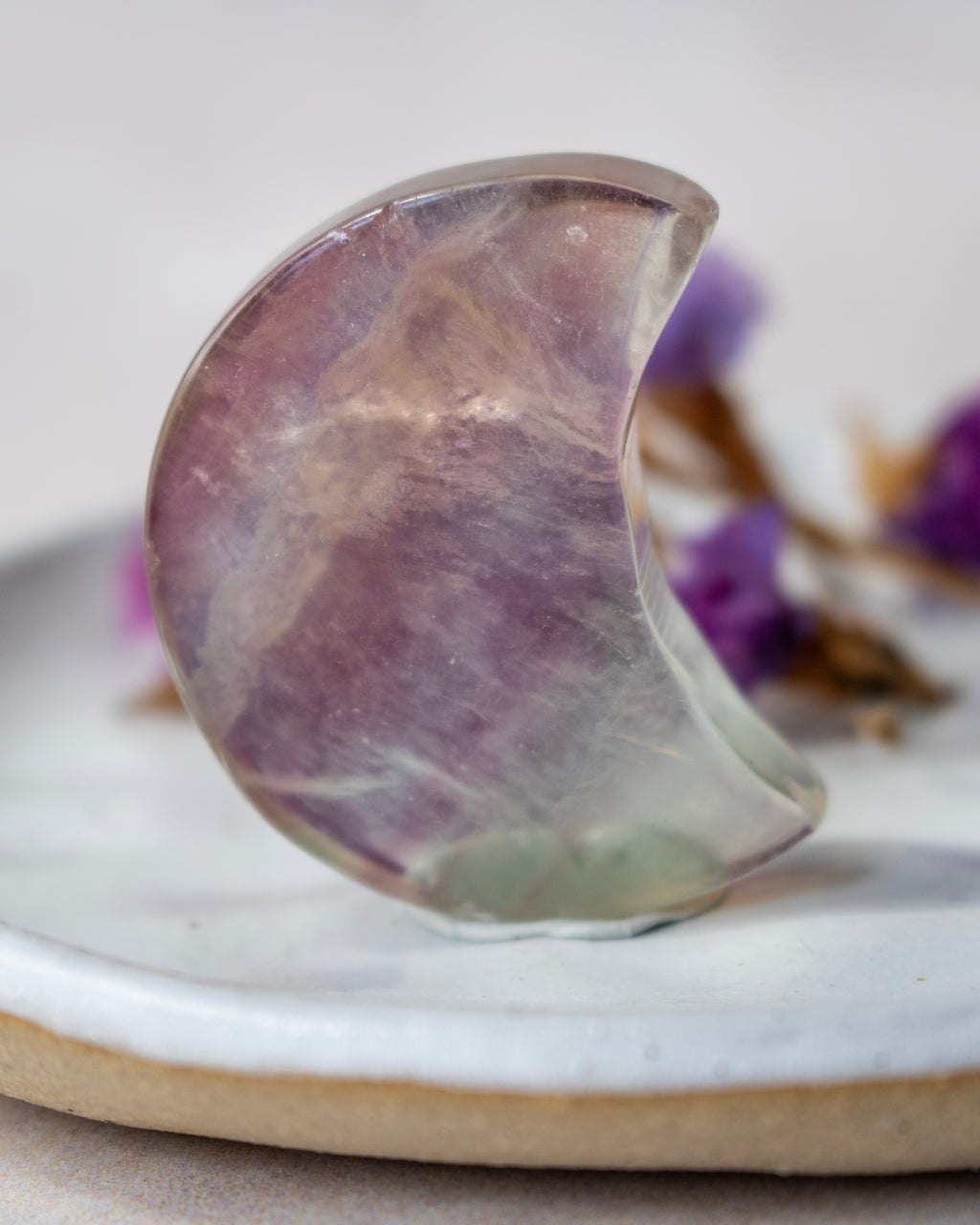 A crescent moon-shaped polished crystal, showing lavender and purple tones with natural internal inclusions, resting on a white ceramic dish with blurred purple flowers in the background.