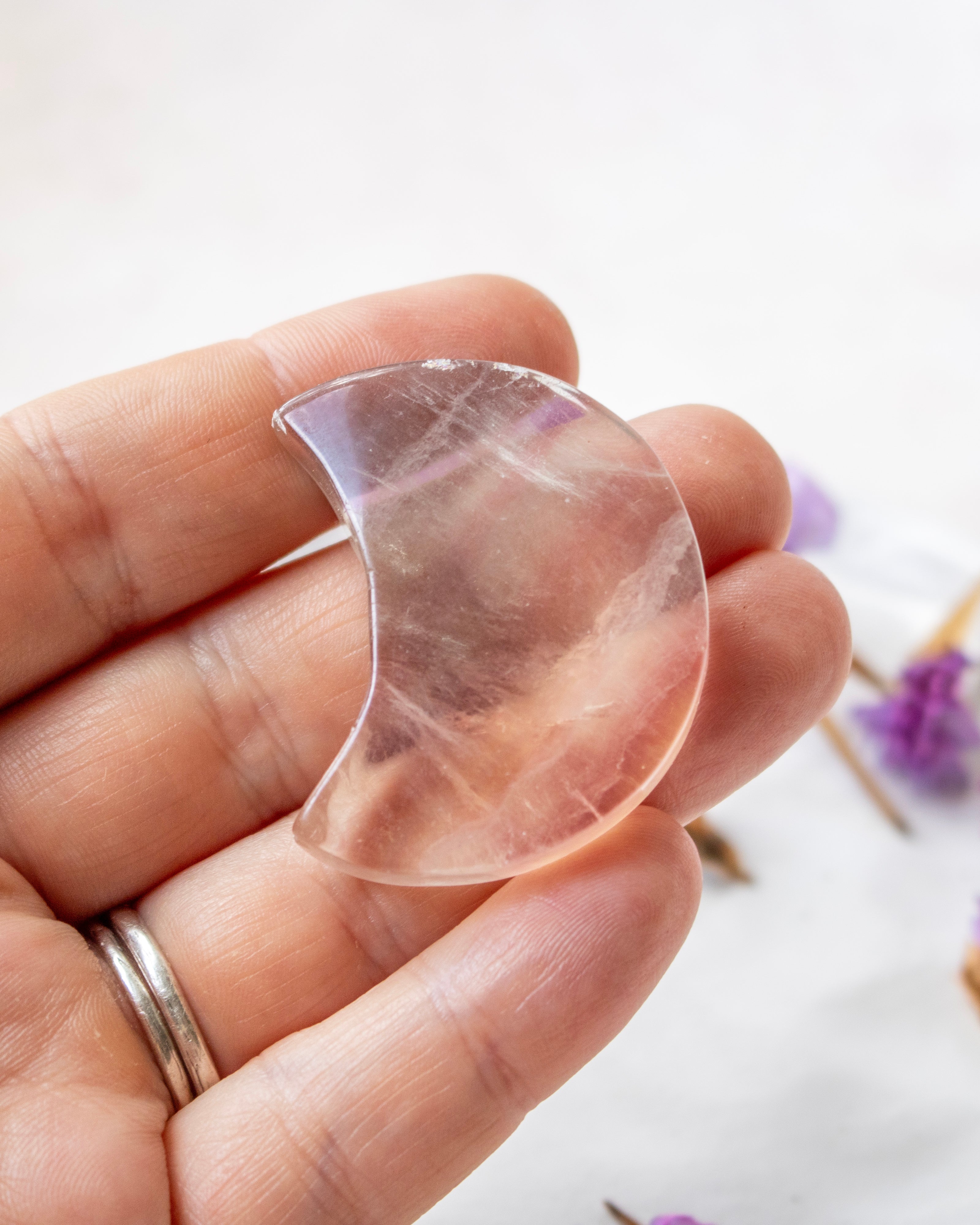 A hand holding a crescent moon-shaped clear crystal pendant against a soft white background with blurred purple flowers.