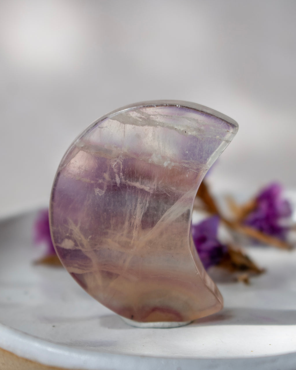 A crescent-shaped translucent purple crystal stone with natural inclusions, standing upright on a white surface with blurred purple flowers in the background.