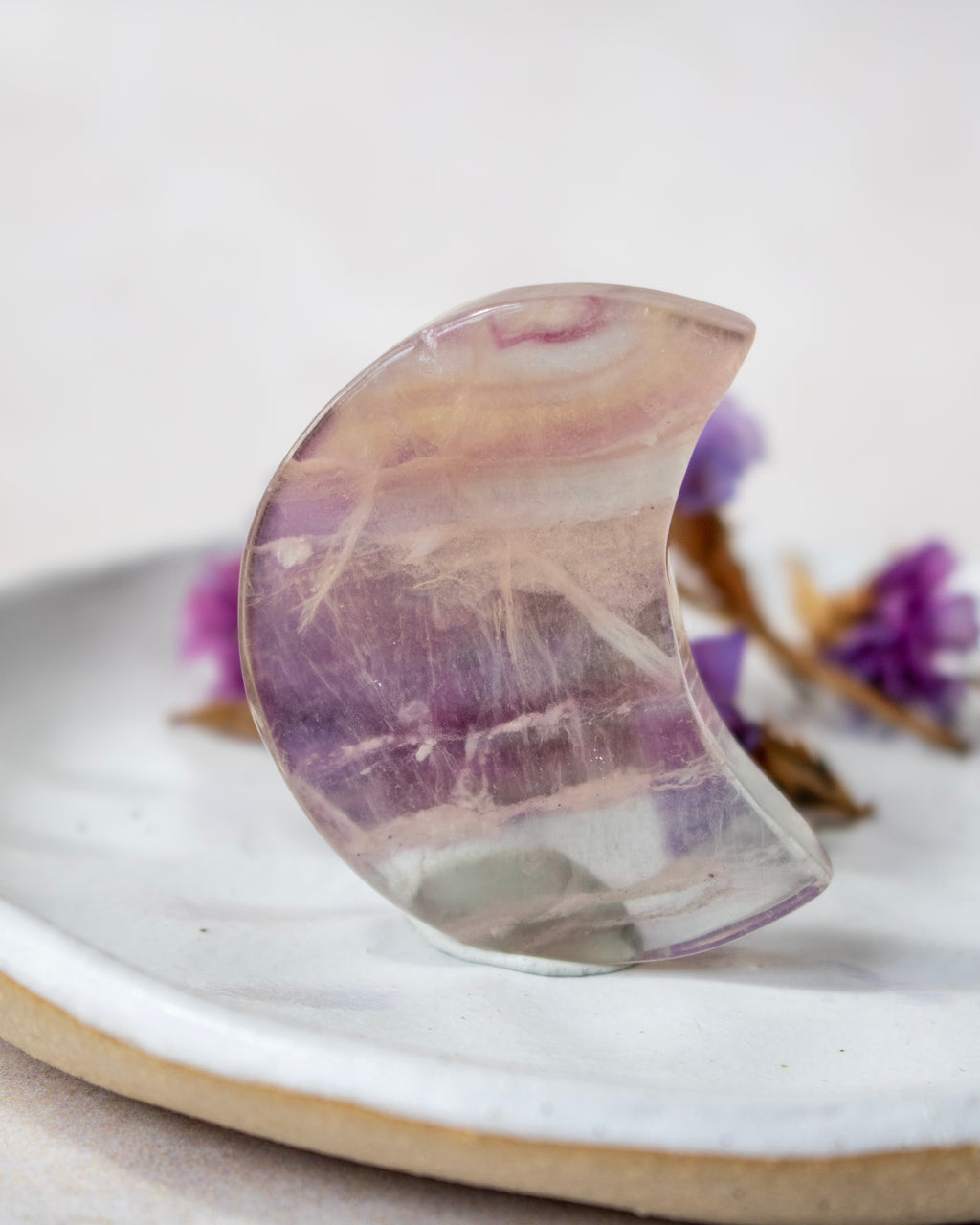 A crescent moon-shaped clear crystal with purple and beige bands, displayed on a white ceramic plate with dried purple flowers in the background.