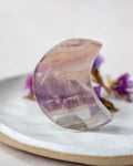 A crescent moon-shaped clear crystal with purple and beige bands, displayed on a white ceramic plate with dried purple flowers in the background.