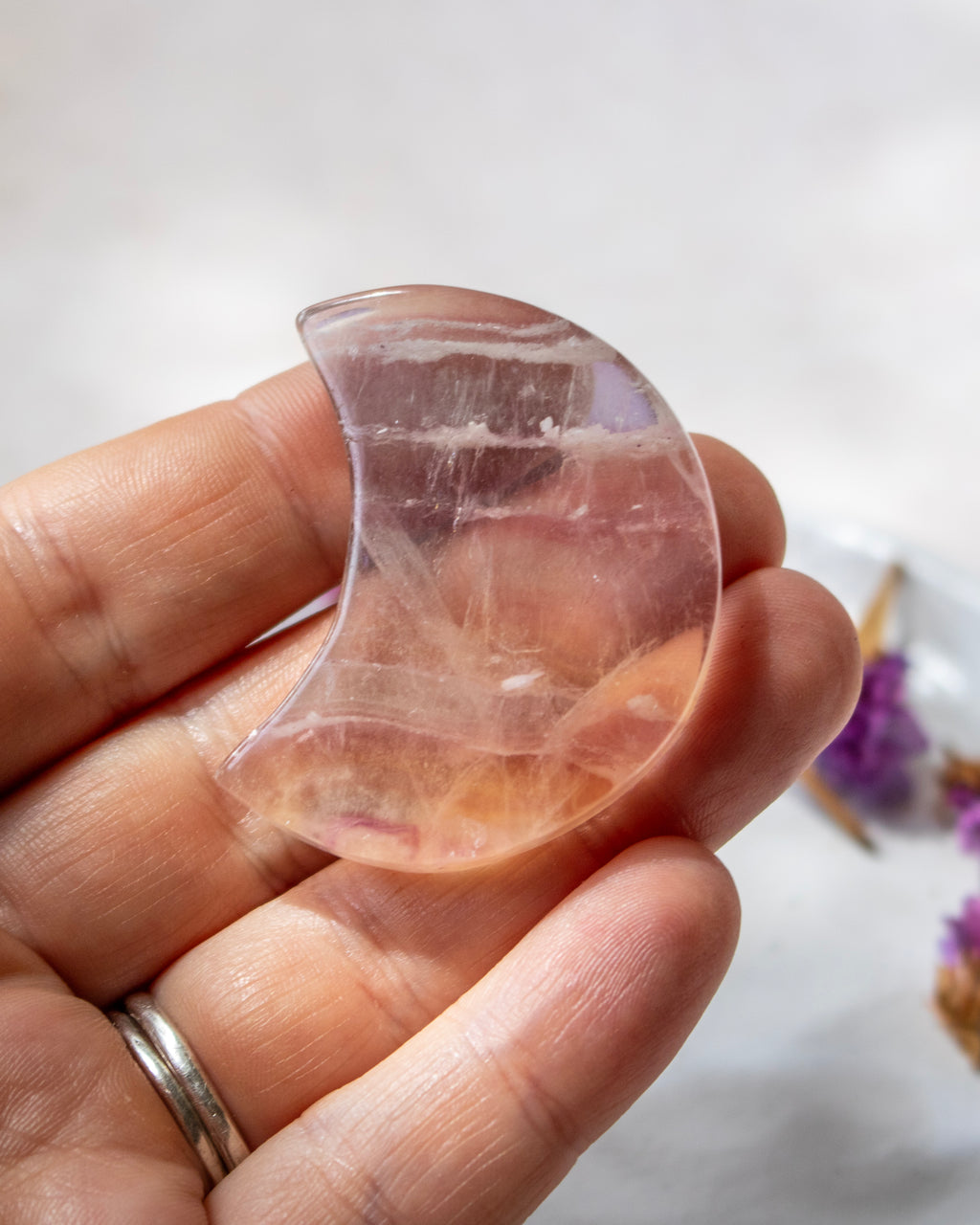 A hand holding a polished crescent moon-shaped clear crystal pendant with natural internal inclusions, shown against a soft white background with blurred purple flowers.