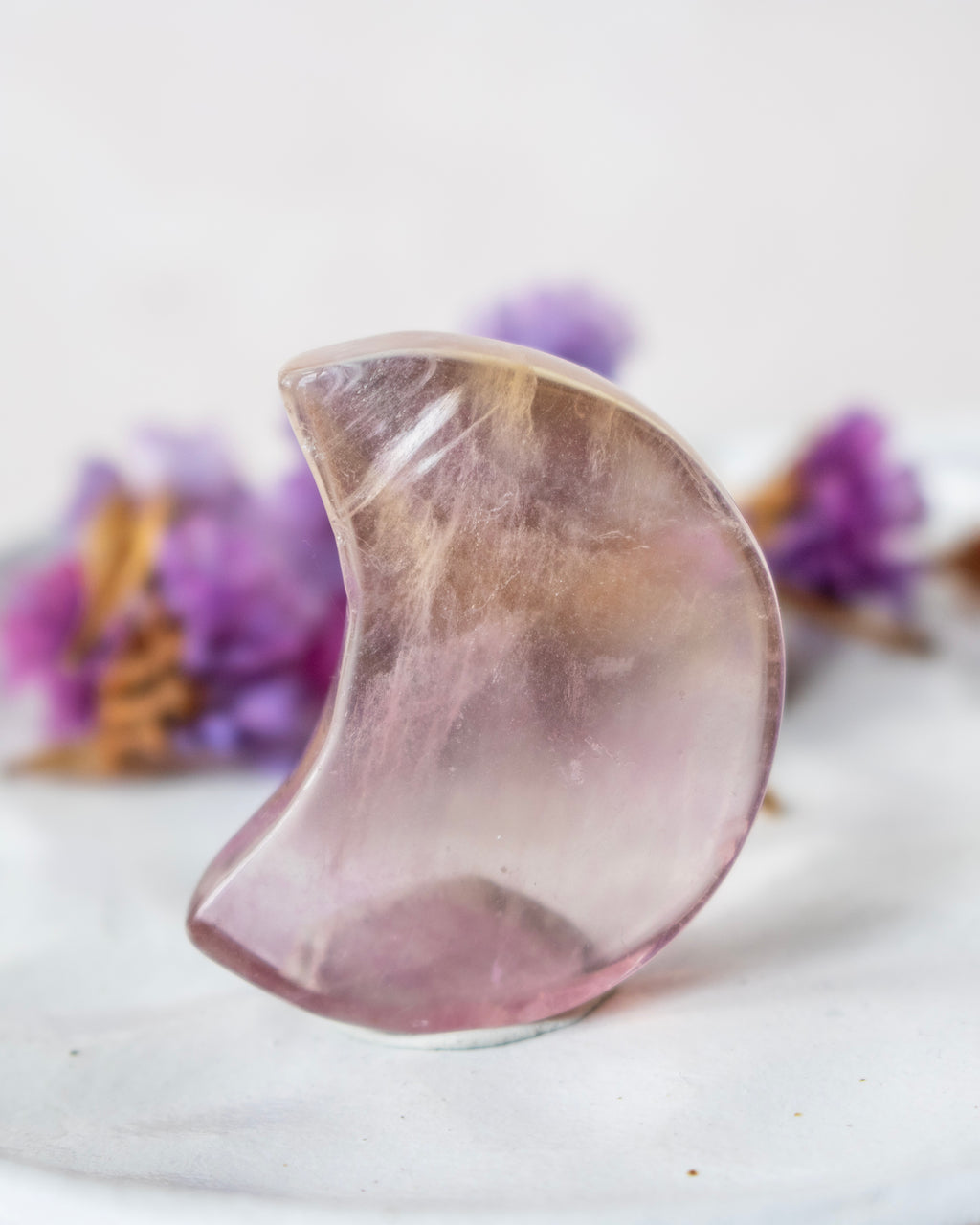 A crescent moon-shaped polished crystal or stone ornament with soft pink and lavender tones, standing upright on a light surface with blurred purple flowers in the background.