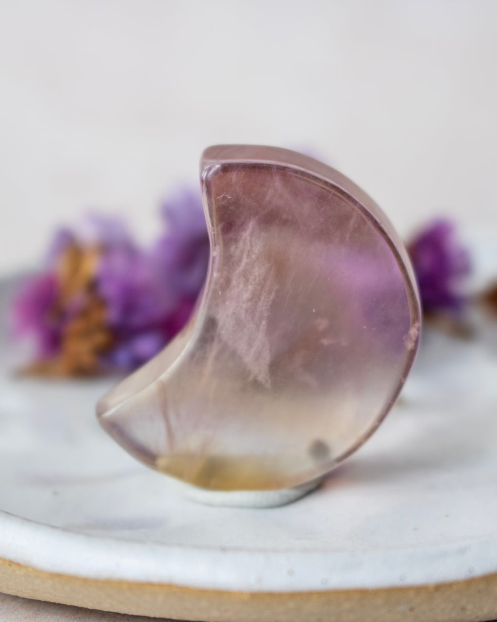A crescent moon-shaped translucent crystal ornament in a soft lavender-purple and smoky gray color, resting on a white ceramic dish with blurred purple flowers in the background.