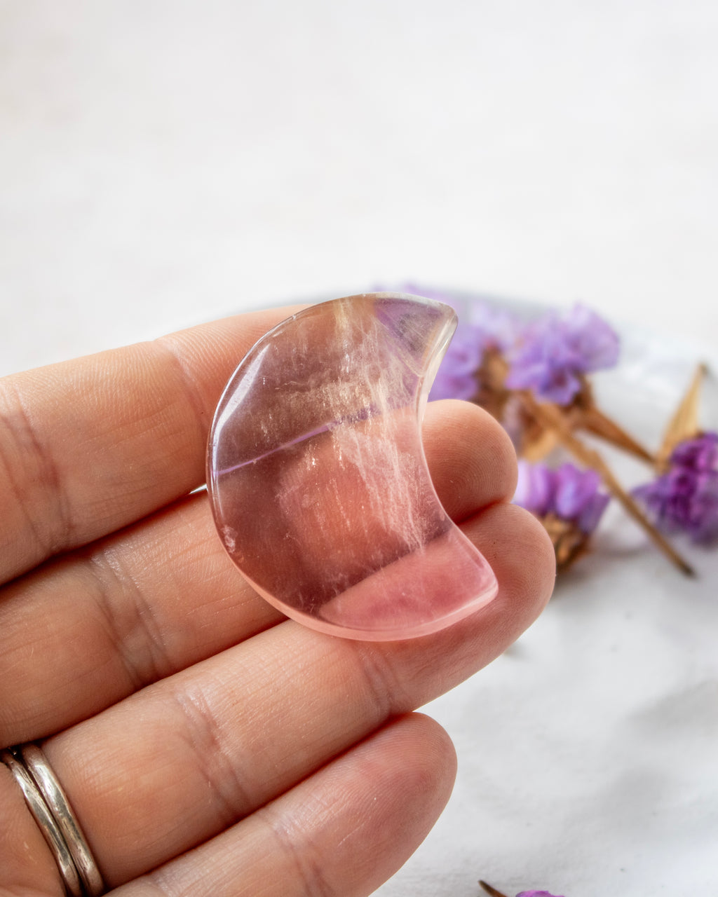 A hand holding a polished crescent moon-shaped crystal pendant, with dried purple flowers blurred in the background.