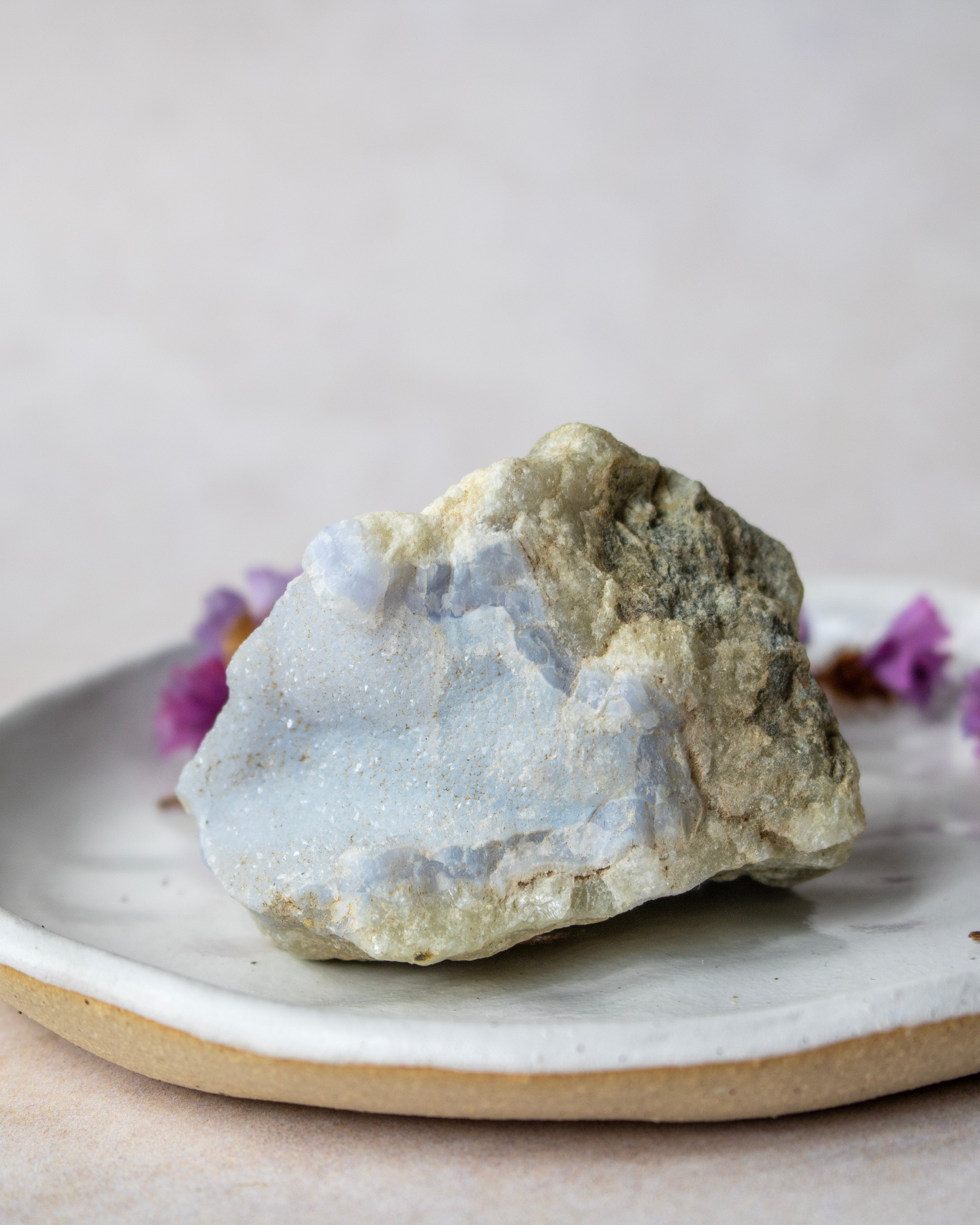 A rough natural crystal or mineral specimen with a pale blue and off-white crystalline surface and tan-gray outer texture, displayed on a ceramic plate against a soft neutral background.