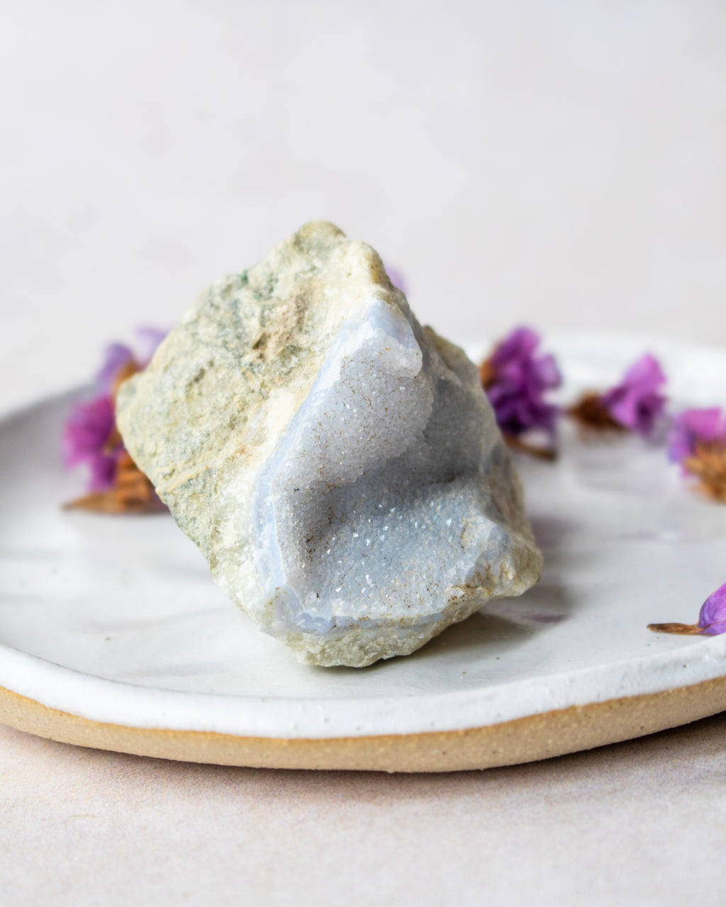 A raw agate crystal cluster with a pale blue geode chamber and light green outer matrix, displayed on a white ceramic plate with dried purple flowers against a soft beige background.