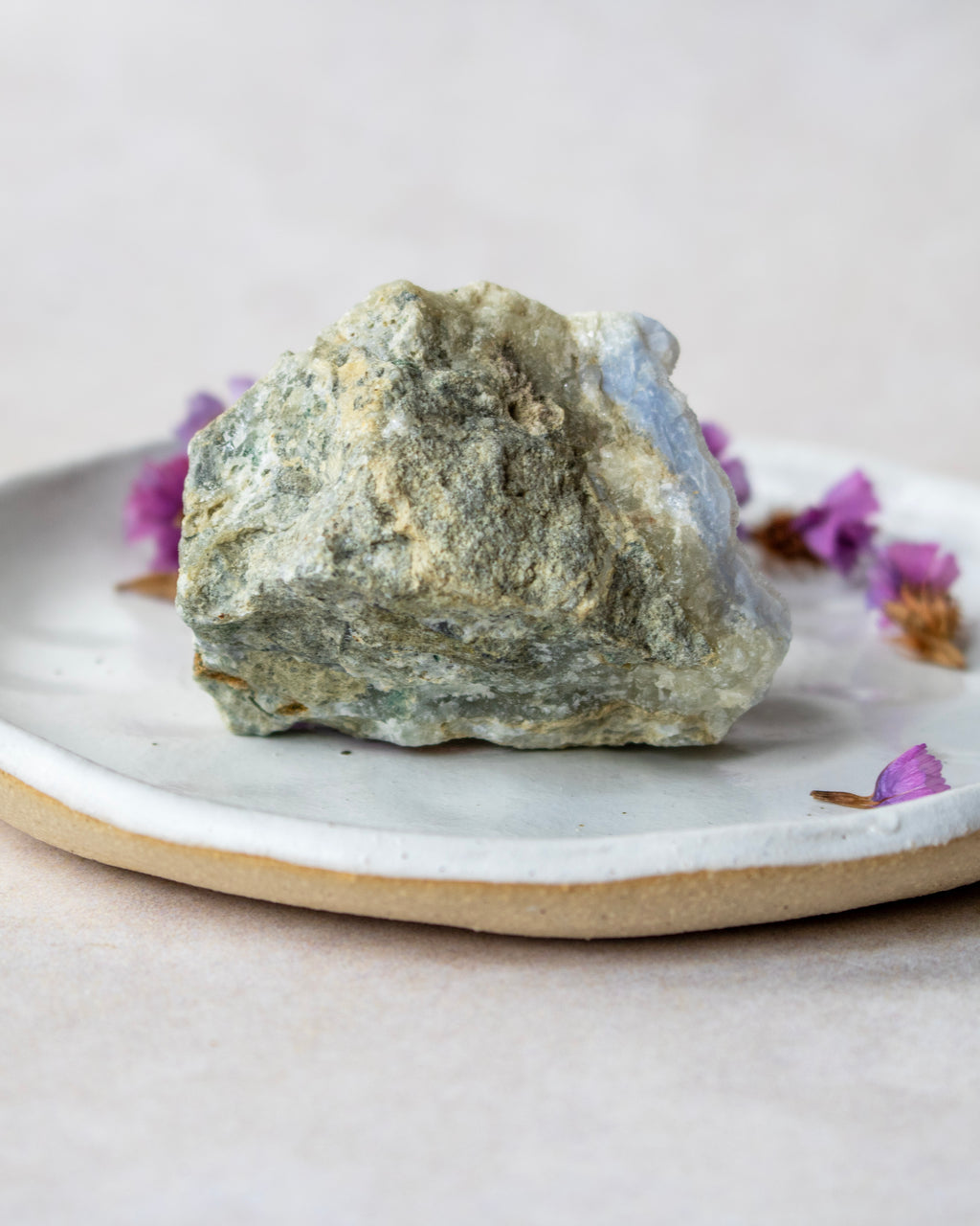 A rough natural stone or crystal with a mottled green, gray, and beige surface sits on a white ceramic plate with a beige rim, styled with small purple dried flowers on a soft neutral background.