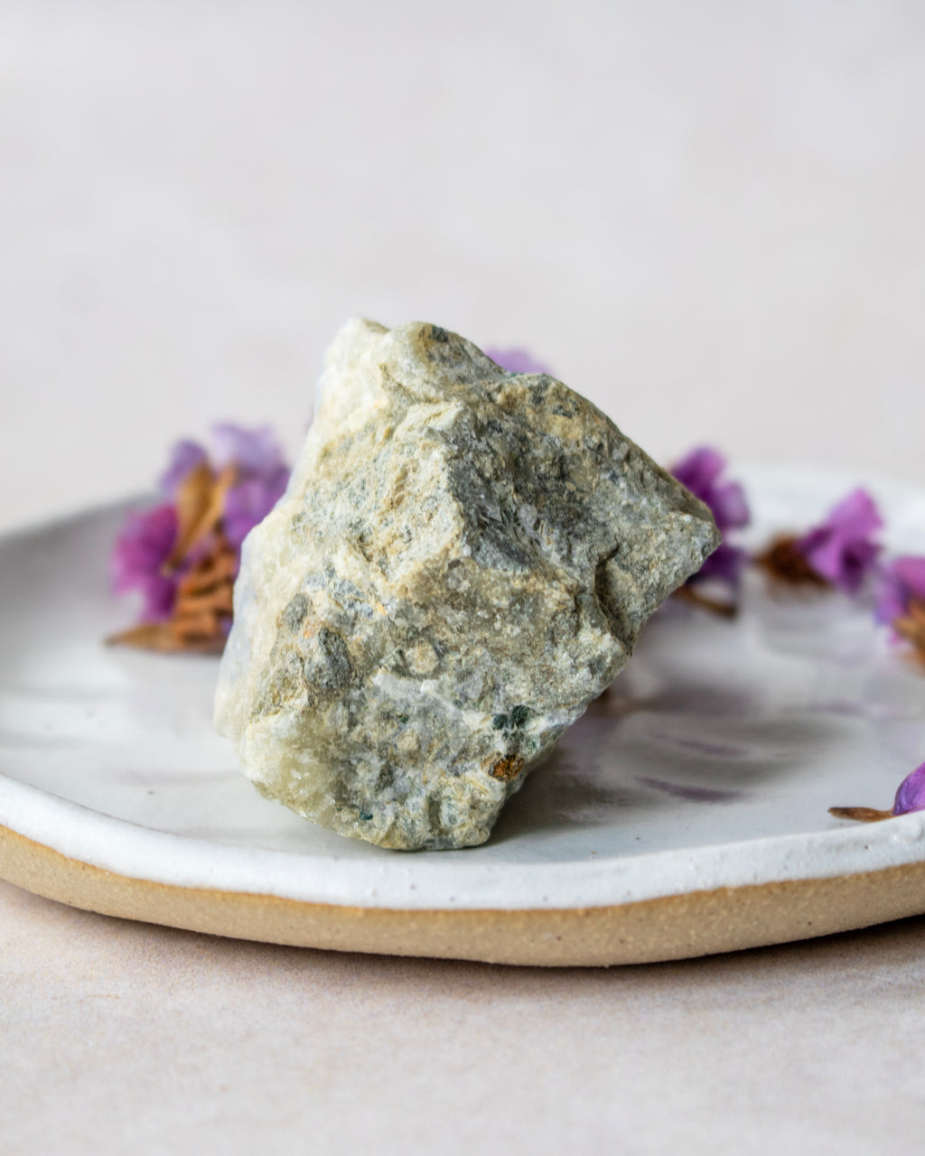A rough, natural stone or crystal with a textured, speckled gray and off-white surface, displayed on a handmade ceramic plate with small purple flowers in the background.