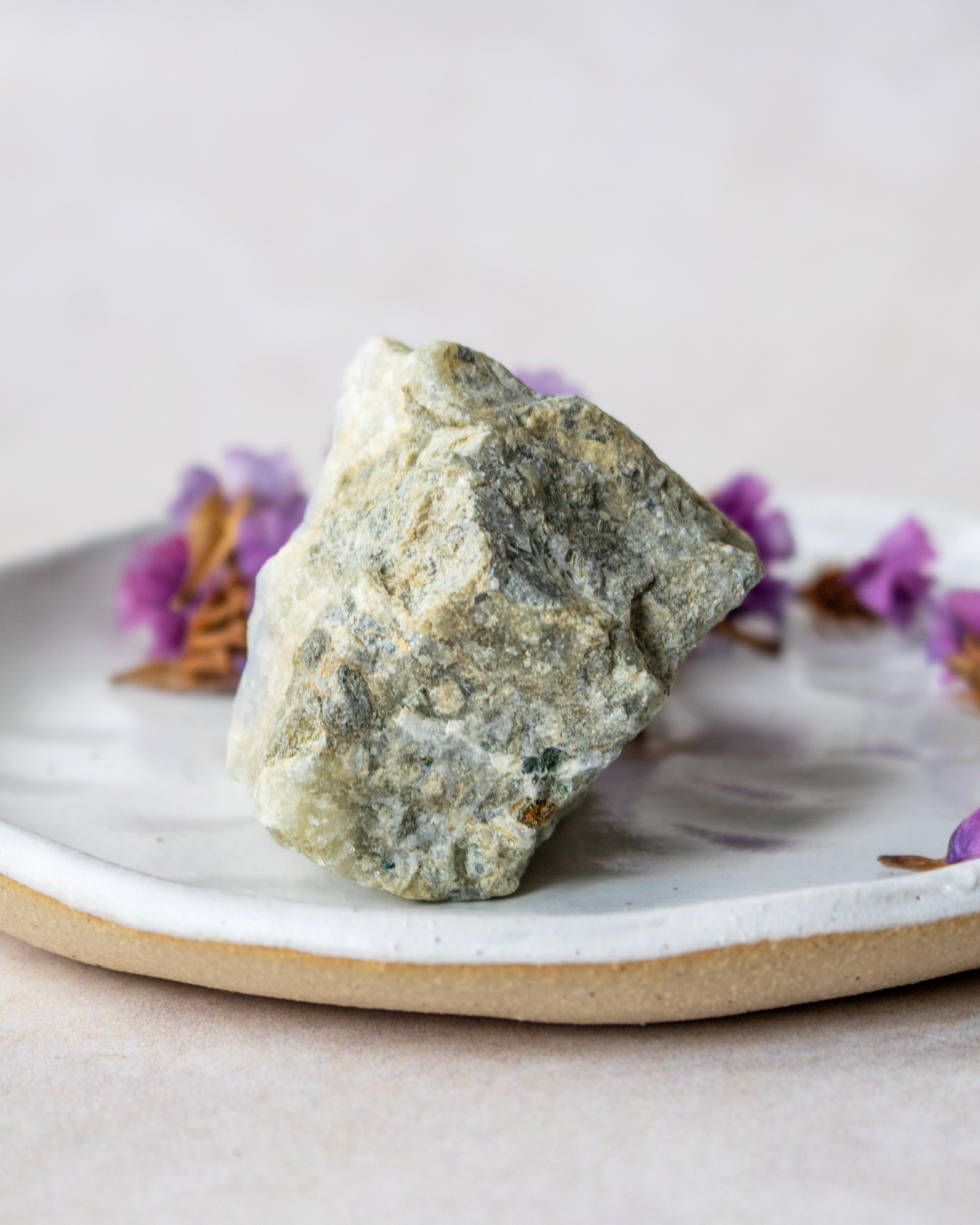 A rough, natural stone or crystal with a textured, speckled gray and off-white surface, displayed on a handmade ceramic plate with small purple flowers in the background.