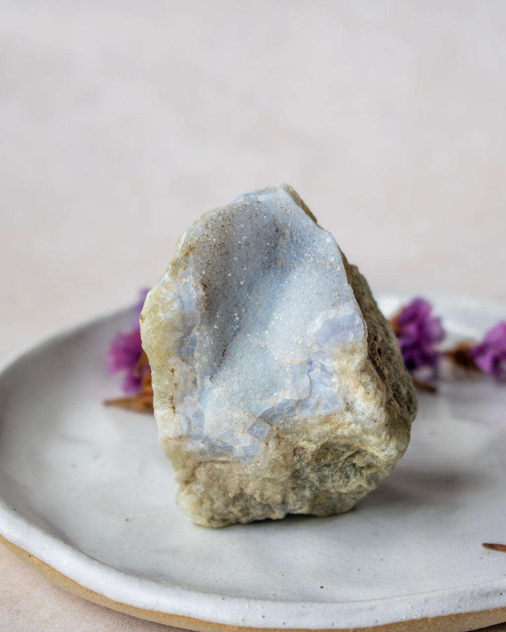 A rough natural crystal or mineral specimen with a pale blue-gray interior and tan outer surface, displayed on a light ceramic plate with small purple flowers in the background.