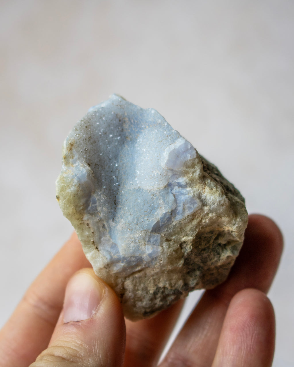 A hand holding a rough natural crystal or mineral specimen with a pale blue-gray druzy-like interior and a brown textured outer matrix, against a soft beige background.