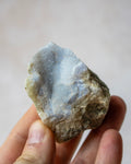 A hand holding a rough natural crystal or mineral specimen with a pale blue-gray druzy-like interior and a brown textured outer matrix, against a soft beige background.
