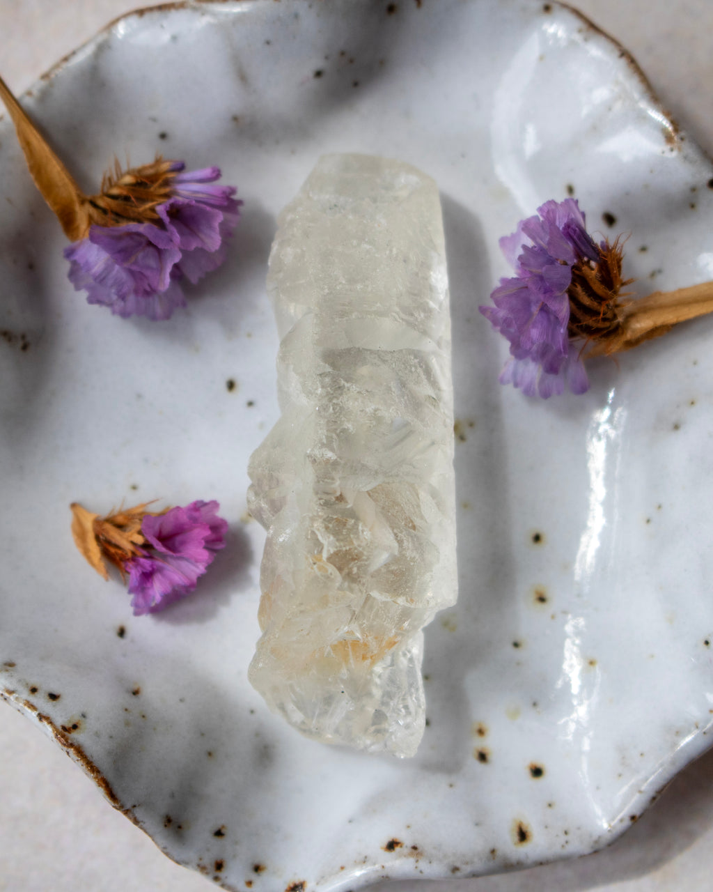 A natural clear quartz crystal cluster sits in the center of a white speckled ceramic dish decorated with dried purple flowers.